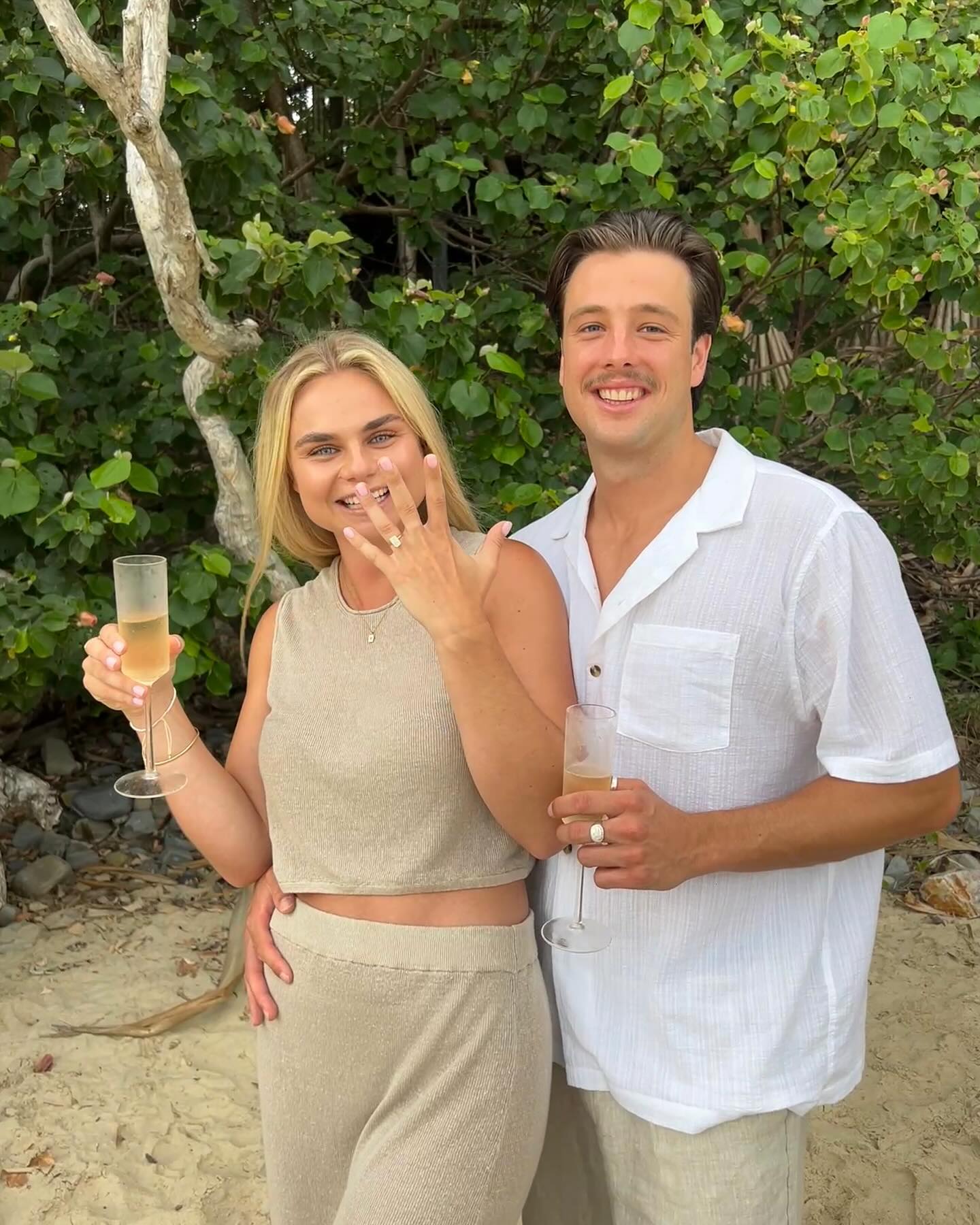 Couple celebrating engagement on a beach, woman showing diamond ring, both holding champagne flutes, with lush greenery in the background.