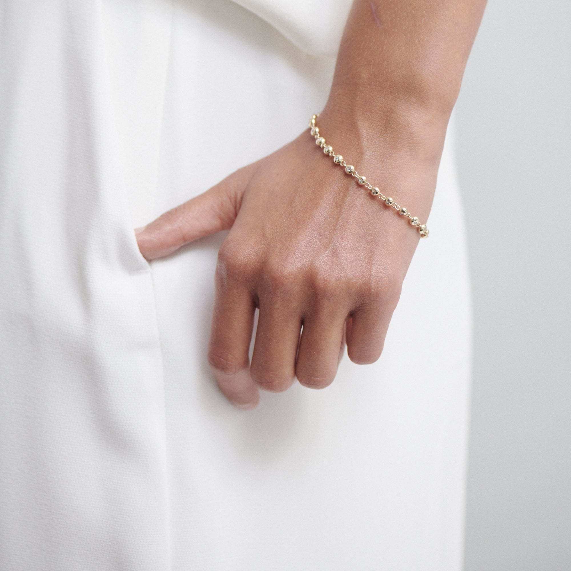 A hand wearing a delicate gold beaded bracelet rests casually in a pocket of an elegant white outfit, showcasing minimalist luxury jewellery.