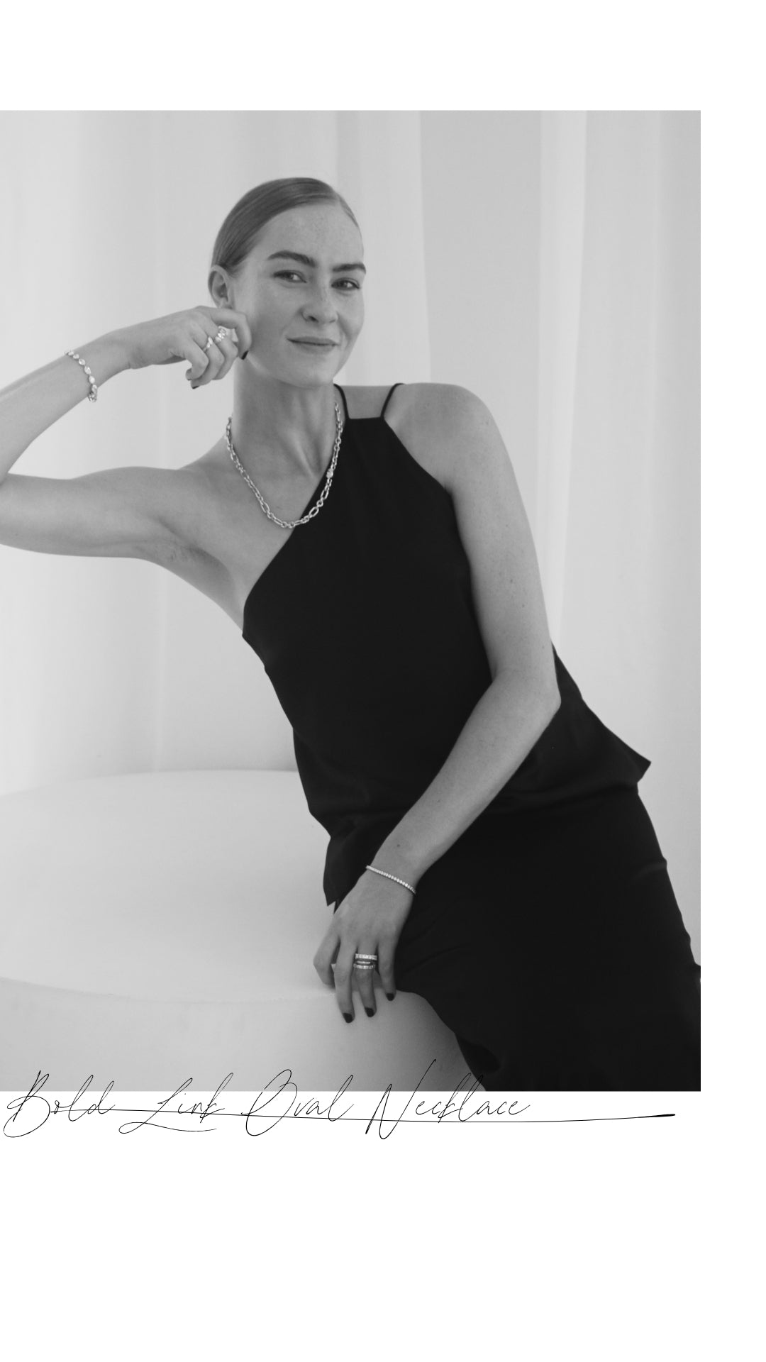 A woman in a black dress wears an elegant oval link necklace. She poses sitting on a white surface, showcasing bracelets and rings.