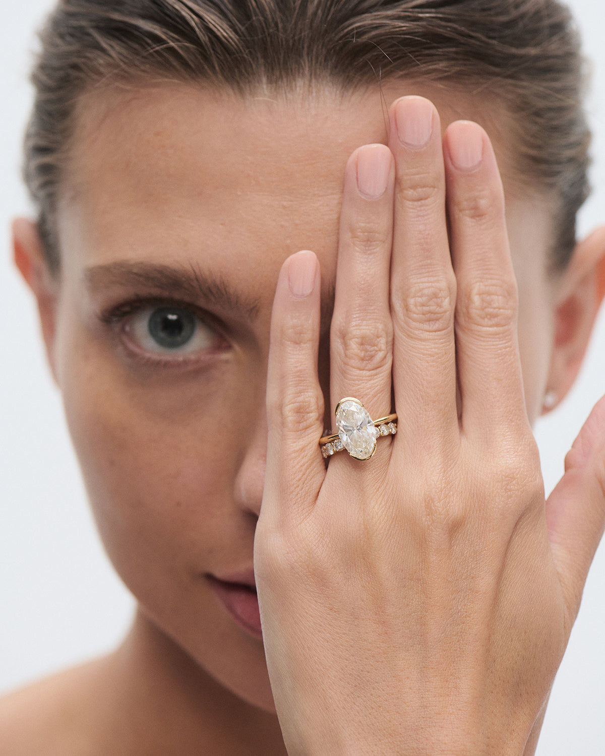 Person with slicked-back hair, covering one eye with a hand adorned with a large, oval-cut diamond ring, showcasing luxury jewellery.