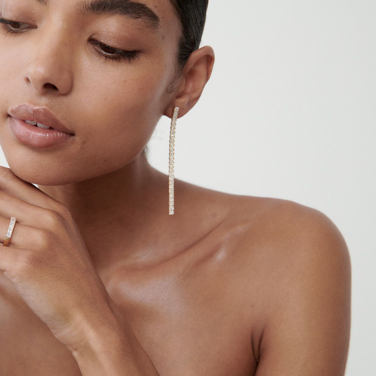Woman wearing long, elegant diamond earrings and matching ring, against a neutral background. Luxury jewellery, minimalistic style.