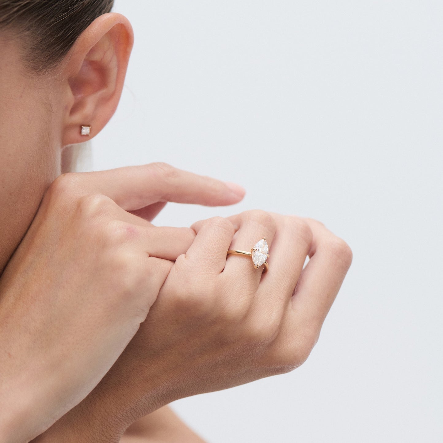 Woman's hand with a marquise-cut diamond ring and a square stud earring. Luxurious jewellery display on light background.