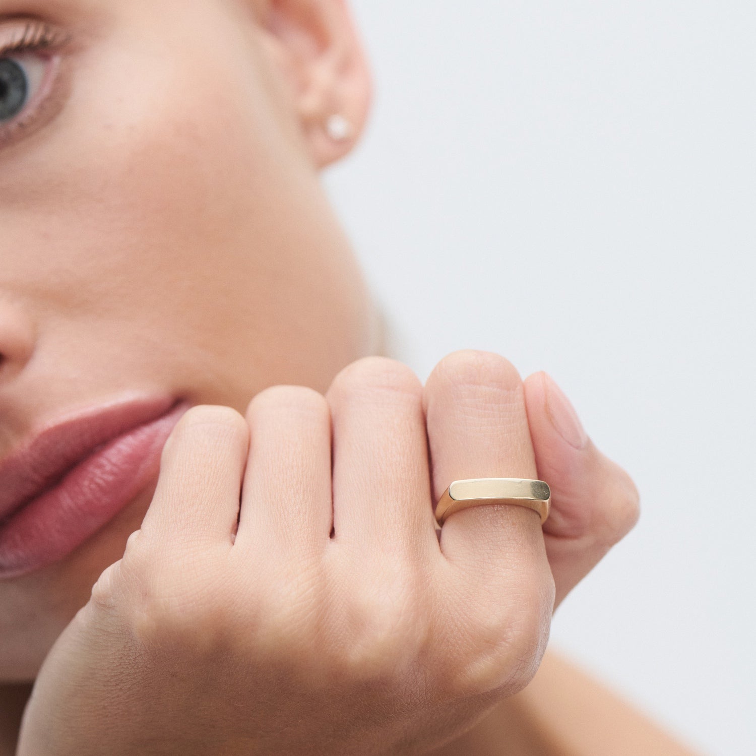 Close-up of a woman's hand wearing a sleek gold band ring, her face slightly visible in the background. Elegant and minimalistic jewellery.