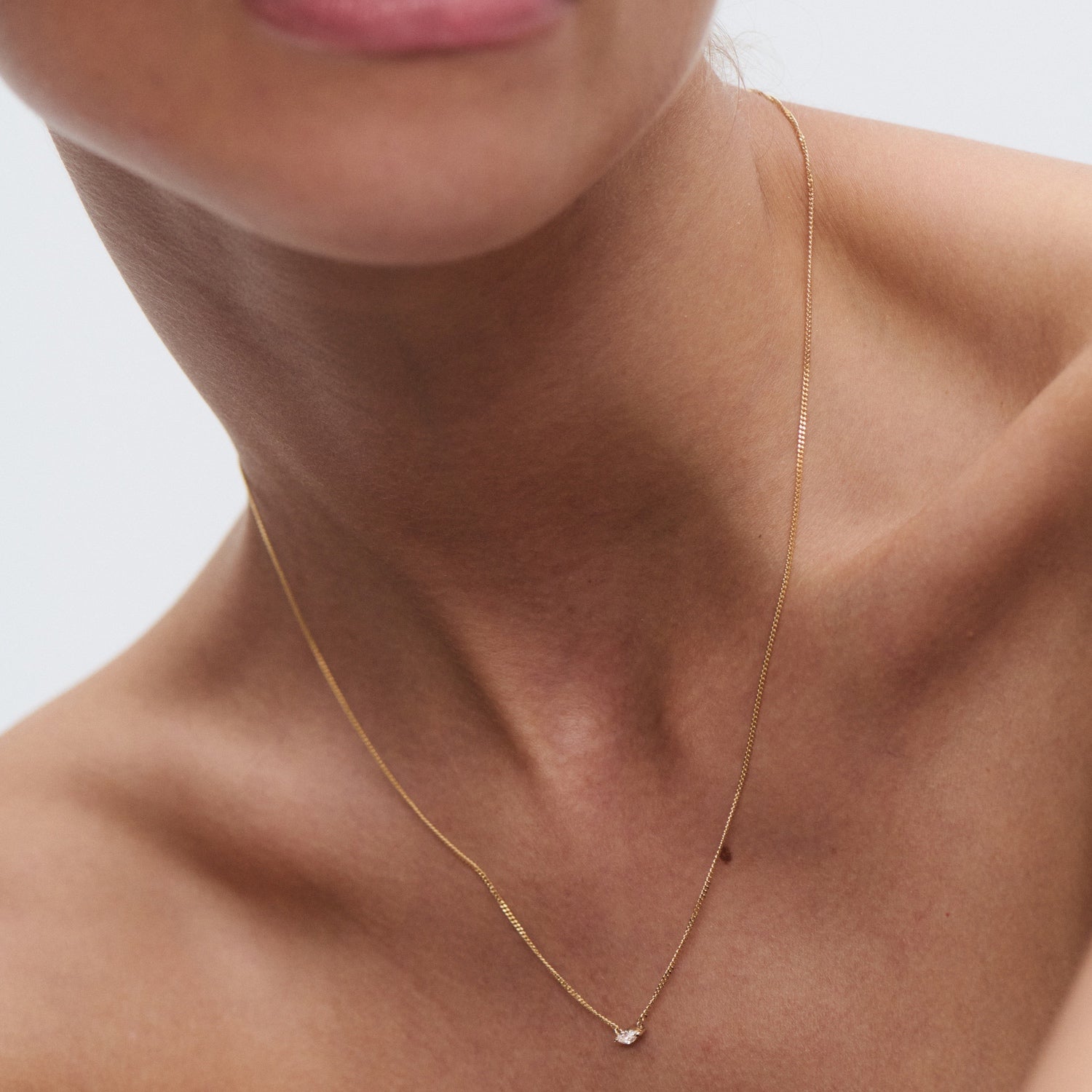 Close-up of a woman's neck wearing a delicate gold chain with a small diamond pendant, against a plain background.