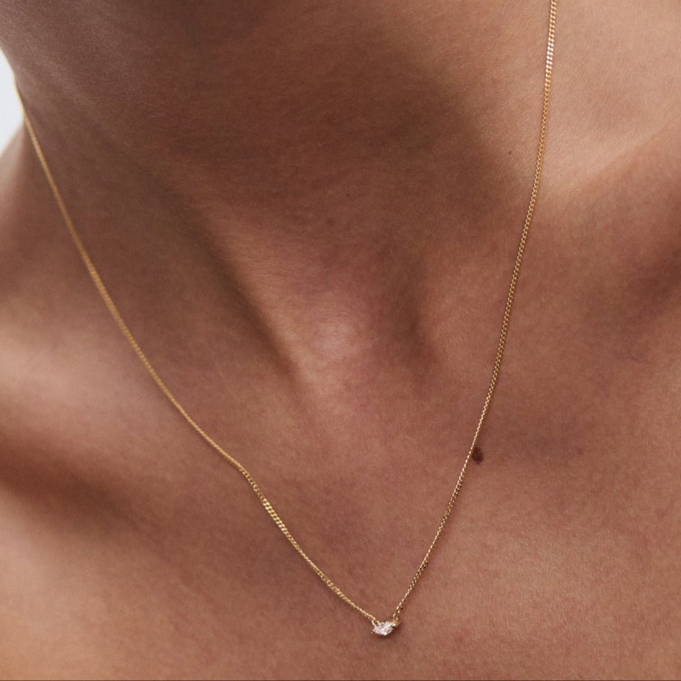 Close-up of a woman's neck wearing a delicate gold chain with a small diamond pendant, against a plain background.