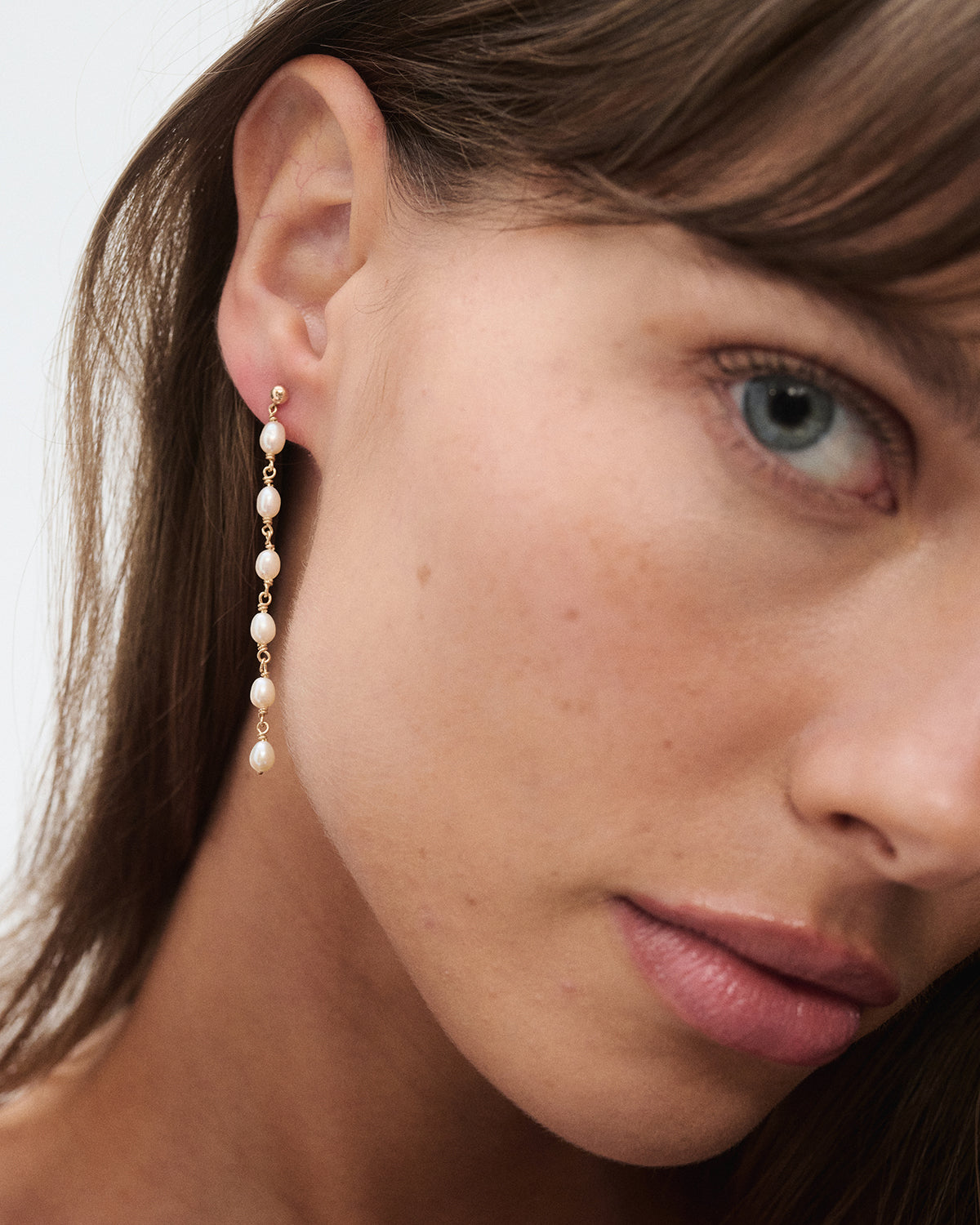 Close-up of a woman's face wearing a long elegant gold earring adorned with small pearls, highlighting luxury jewellery design.