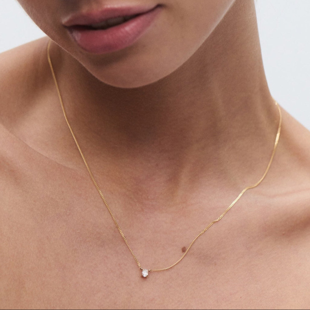 Woman wearing minimalist gold necklace with a small pendant and matching stud earring. Neutral background enhances elegance and luxury. 