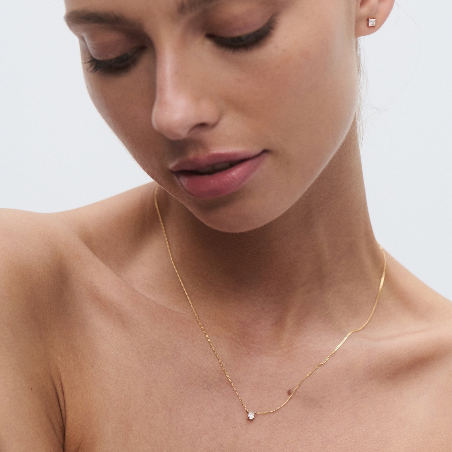 Woman wearing minimalist gold necklace with a small pendant and matching stud earring. Neutral background enhances elegance and luxury.
