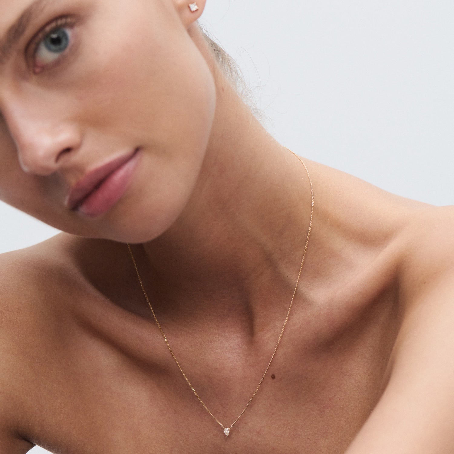 Close-up of a woman wearing a delicate gold necklace with a small diamond pendant and gold earrings, showcasing minimalist luxury jewellery.