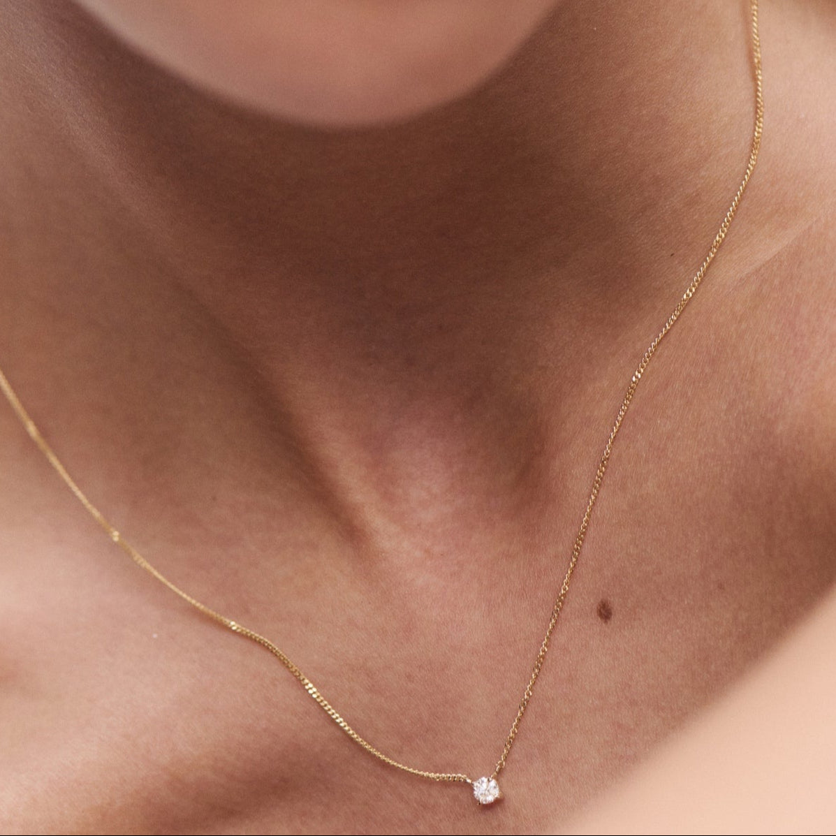 Close-up of a delicate gold necklace with a single diamond pendant, worn by a person with smooth skin. Minimalist luxury jewellery.