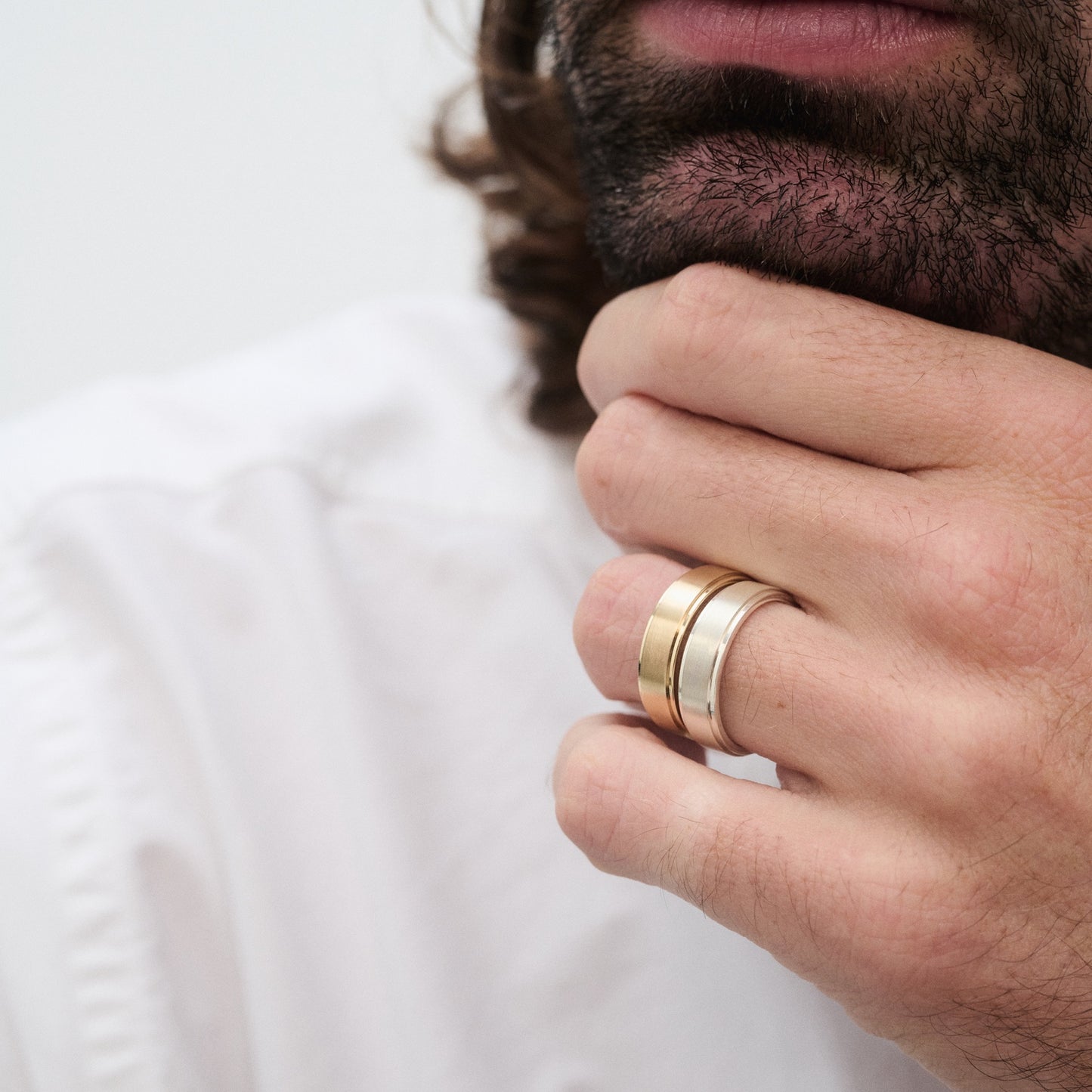 Man wearing three stacked gold and silver rings, resting his chin on his hand, wearing a white shirt. Focus on luxury men's jewellery.