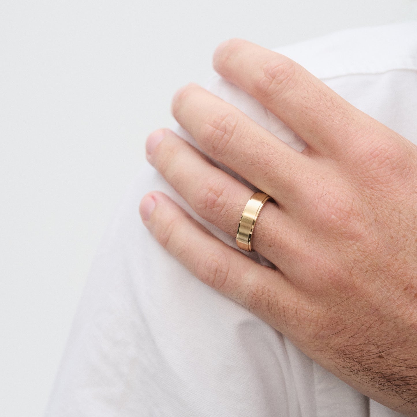 Hand wearing a sleek gold wedding band on the ring finger, resting on a white shirt. Elegant luxury jewellery.