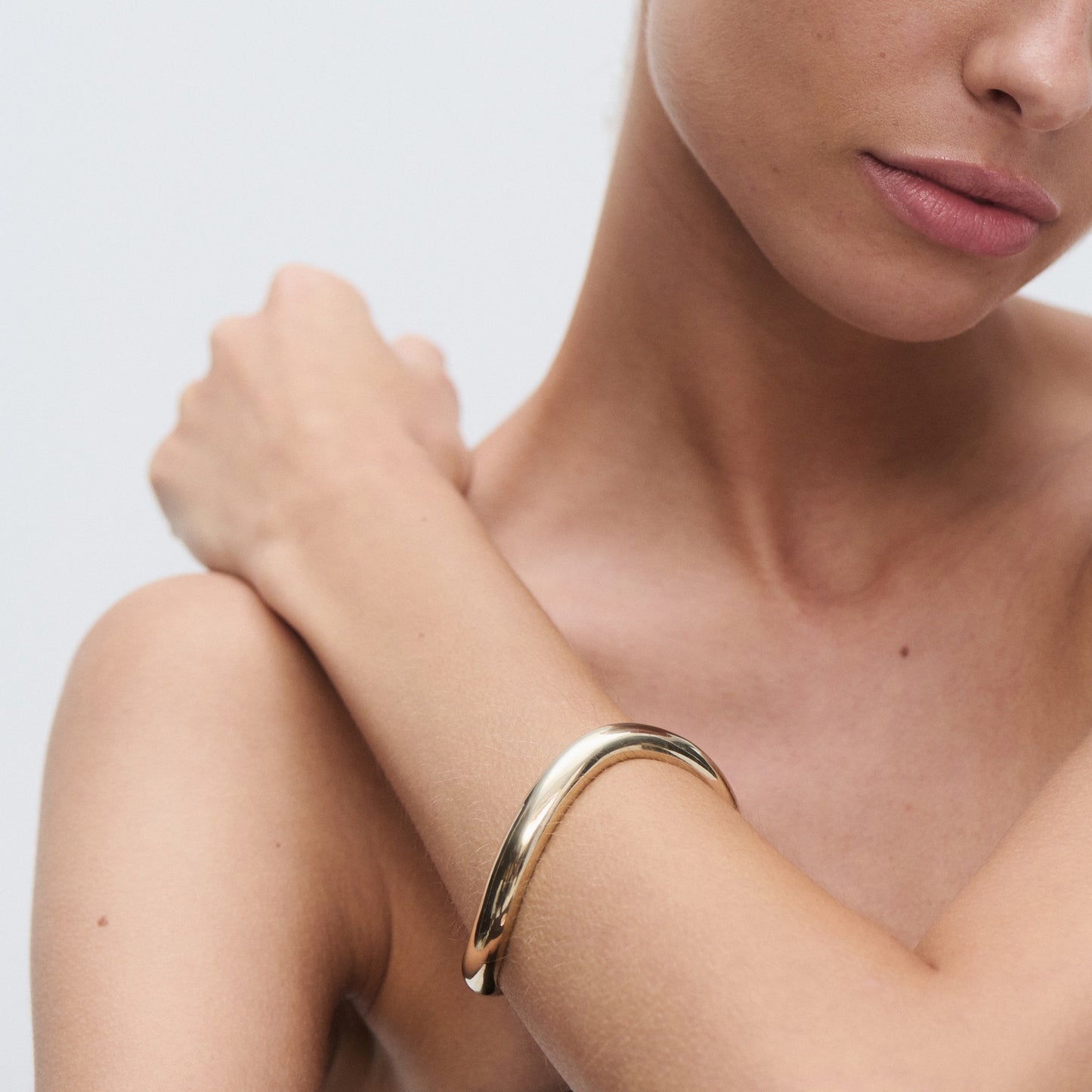 Woman's shoulder and arm with a sleek, polished gold bangle bracelet, showcasing minimalist luxury jewellery. Light background.