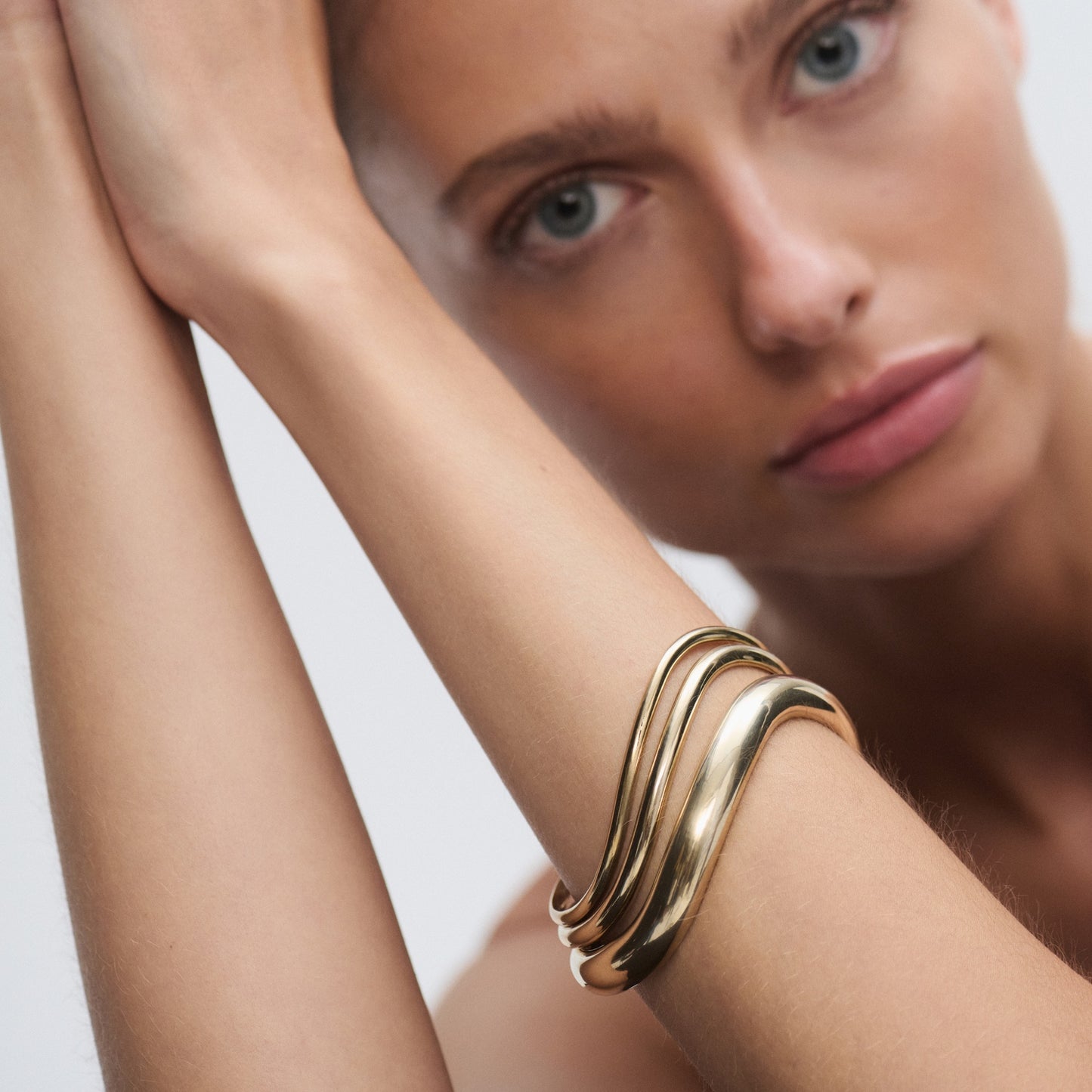 Model wearing an elegant, wavy gold bracelet; luxury jewellery close-up. Pale blue eyes, focused expression, soft background.