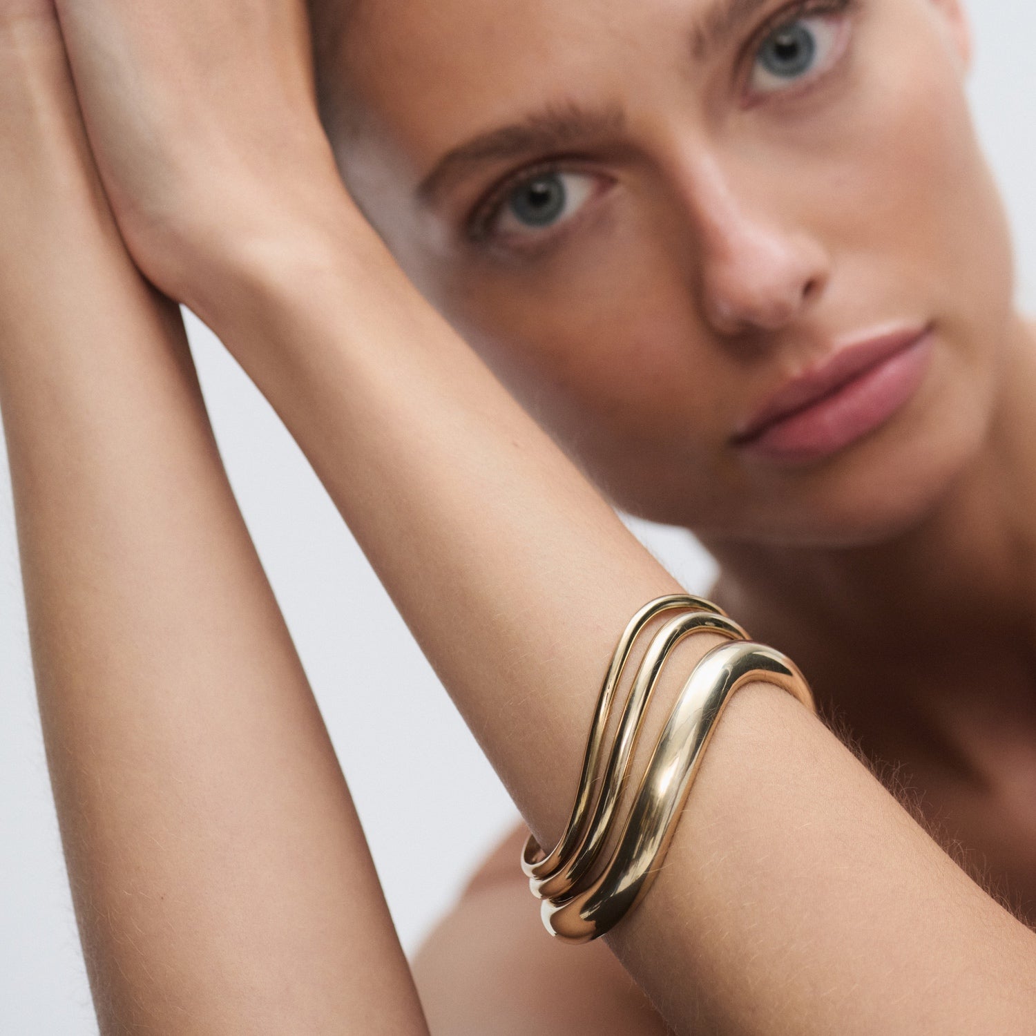 Model wearing an elegant, wavy gold bracelet; luxury jewellery close-up. Pale blue eyes, focused expression, soft background.