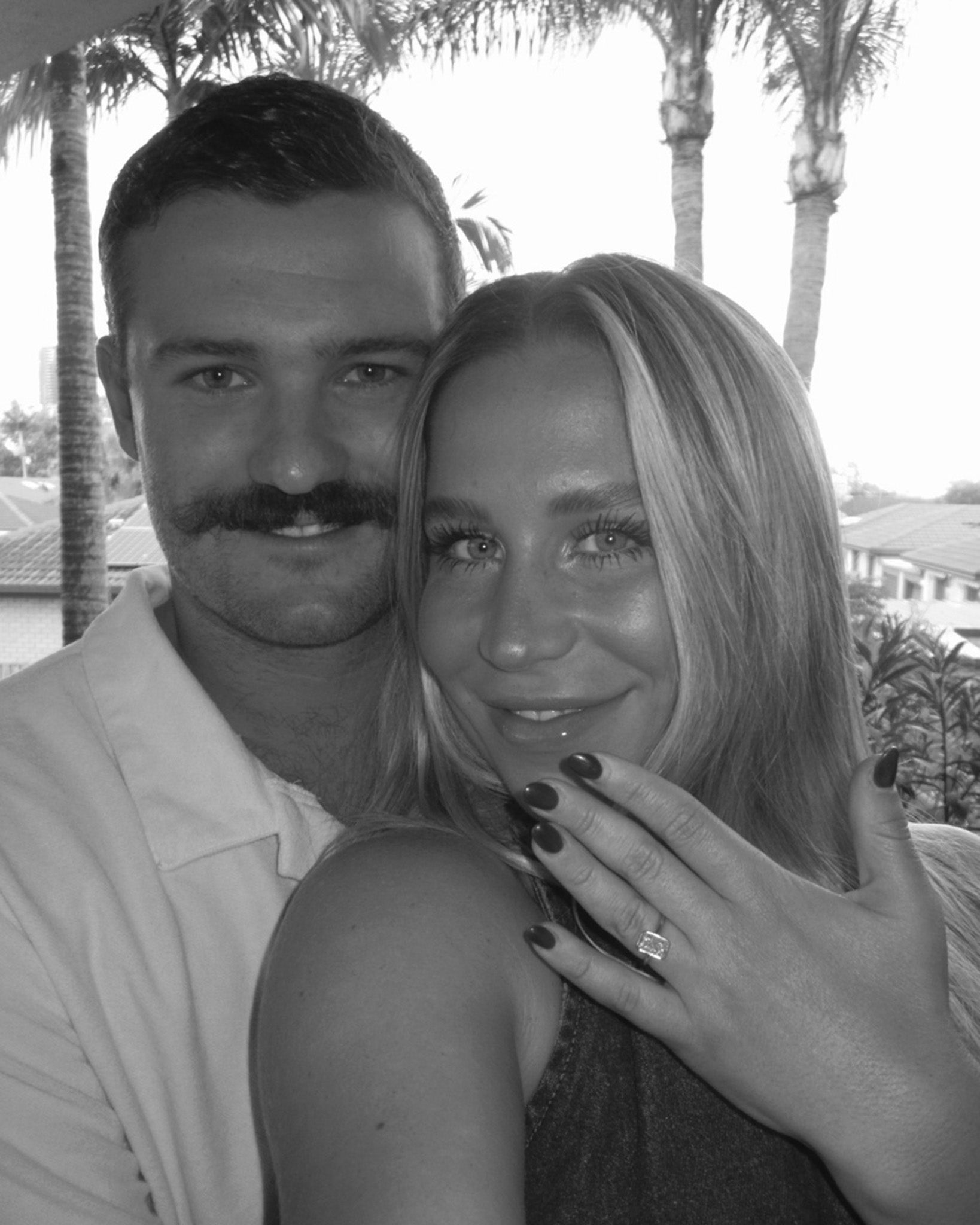 A woman displays an elegant engagement ring with a sparkling diamond, while smiling with a man in a tropical setting. Palms are visible in the background.