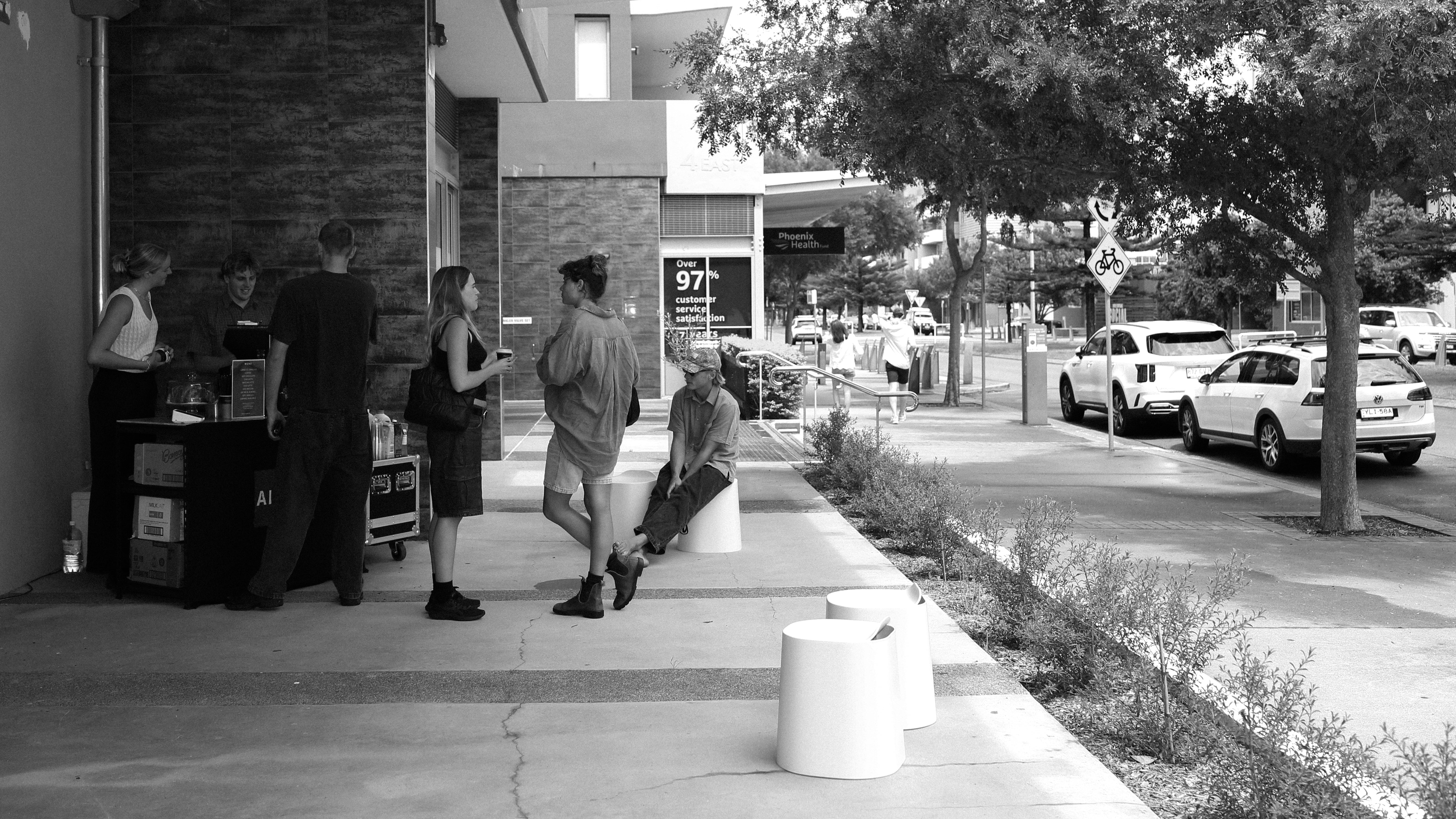 Street scene with people gathered outside a modern building, cars parked along a tree-lined road. Urban, contemporary, social interaction.