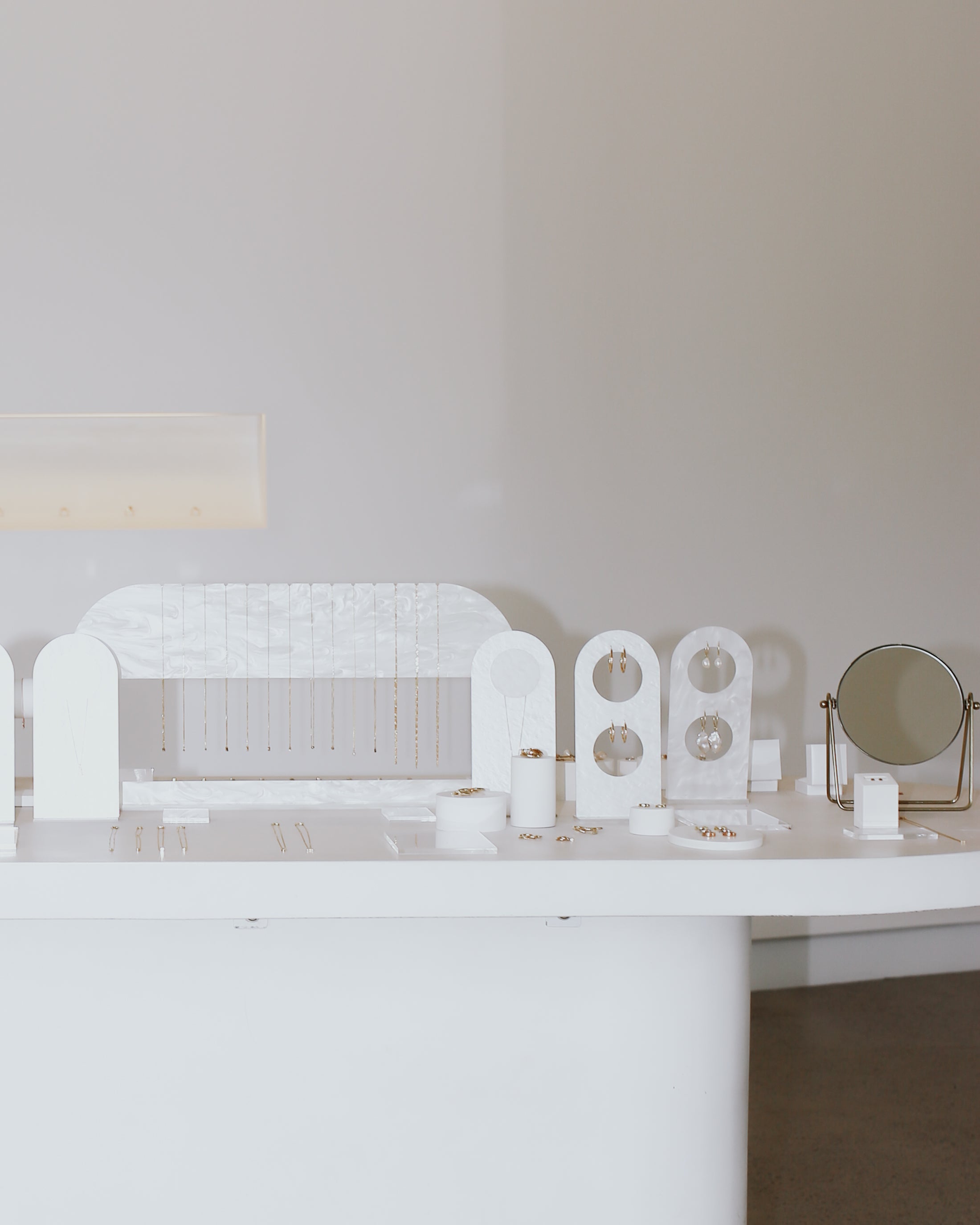 Minimalist luxury jewellery display with white stands, featuring earrings and delicate necklaces, next to a small round mirror on a sleek table.