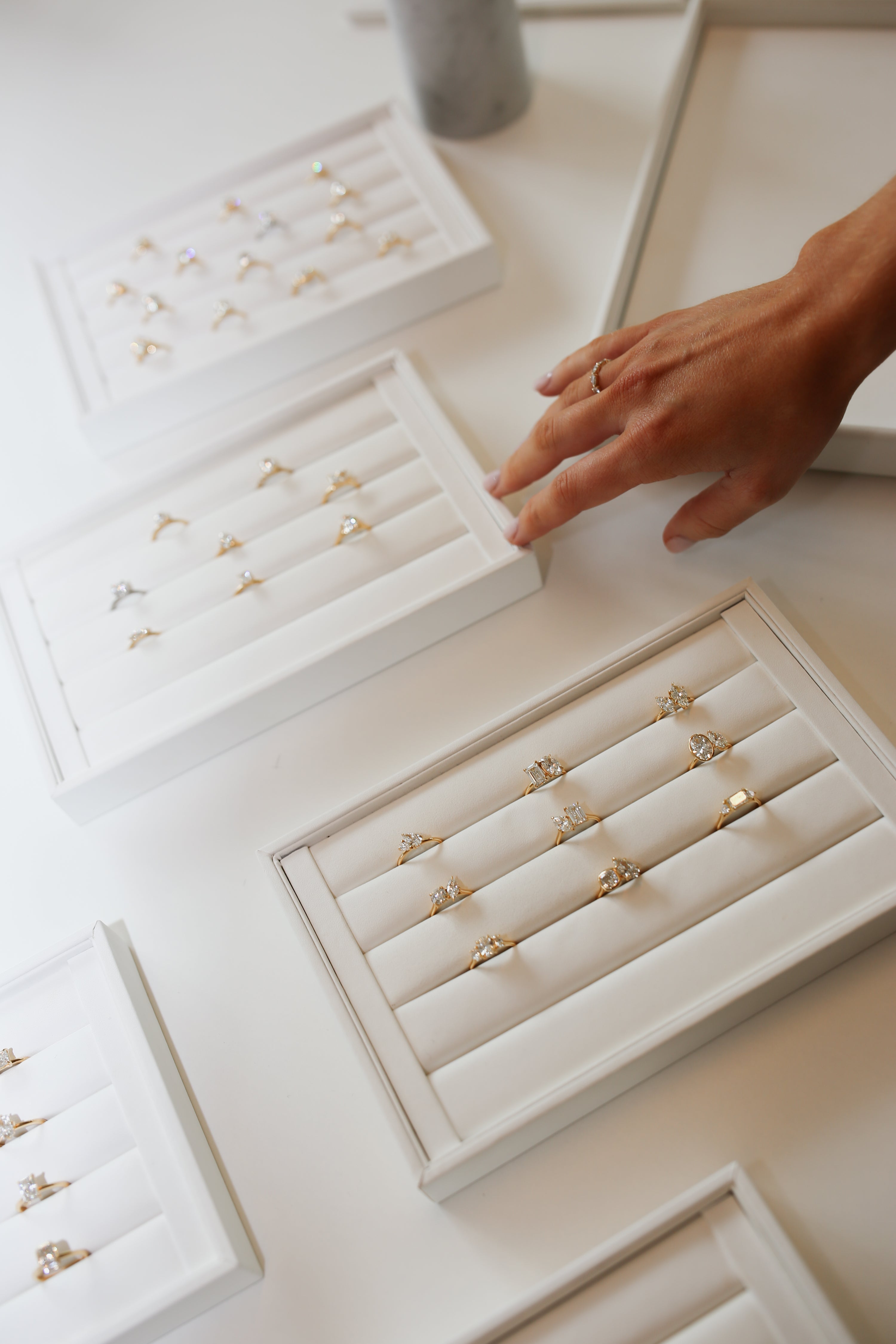 Elegant gold and diamond rings displayed in white jewellery trays, with a hand reaching to select one. High-end jewellery shopping scene.