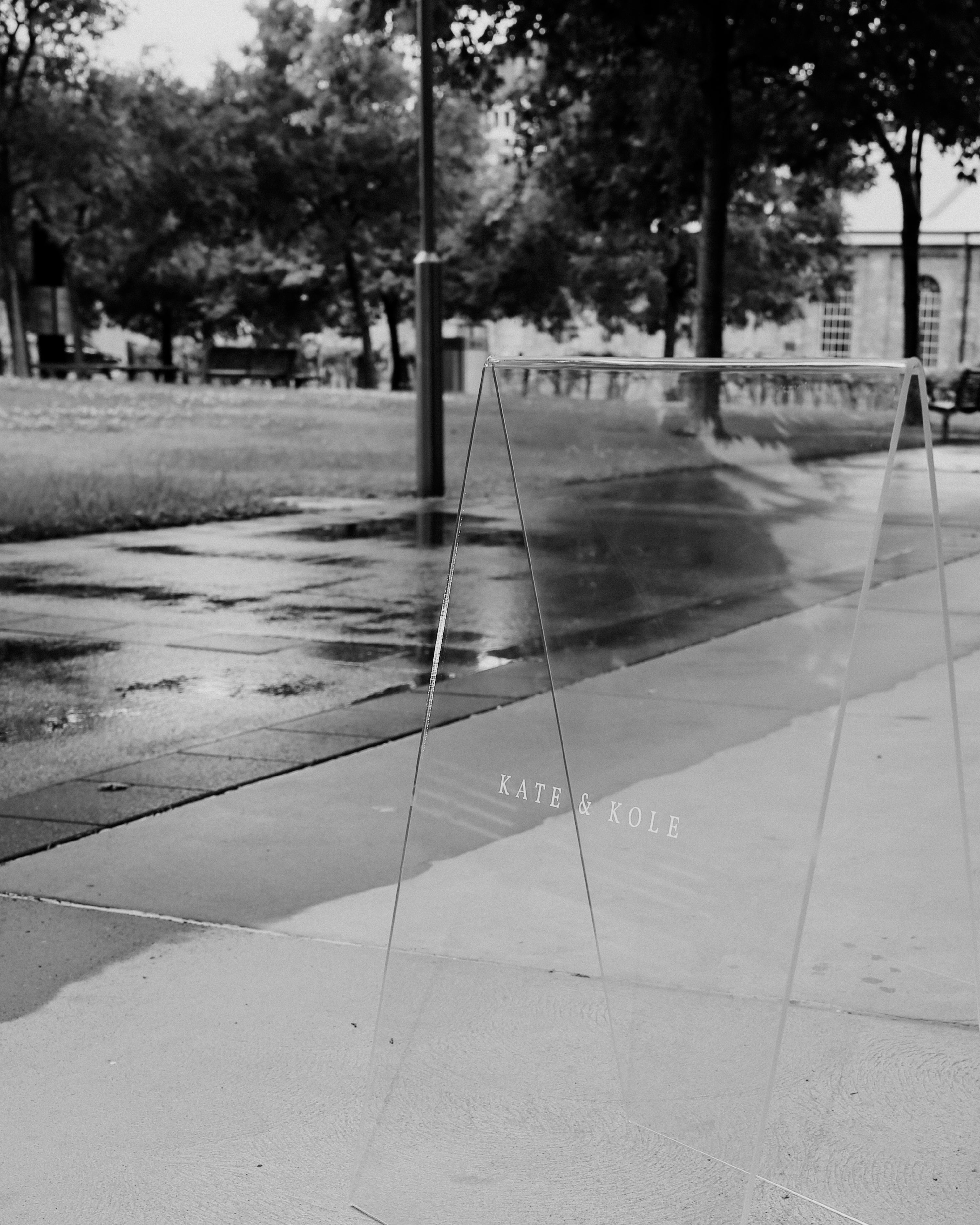 Transparent sign with "Kate & Kole" on a wet pavement, surrounded by trees and grass in an urban park setting. Overcast day.