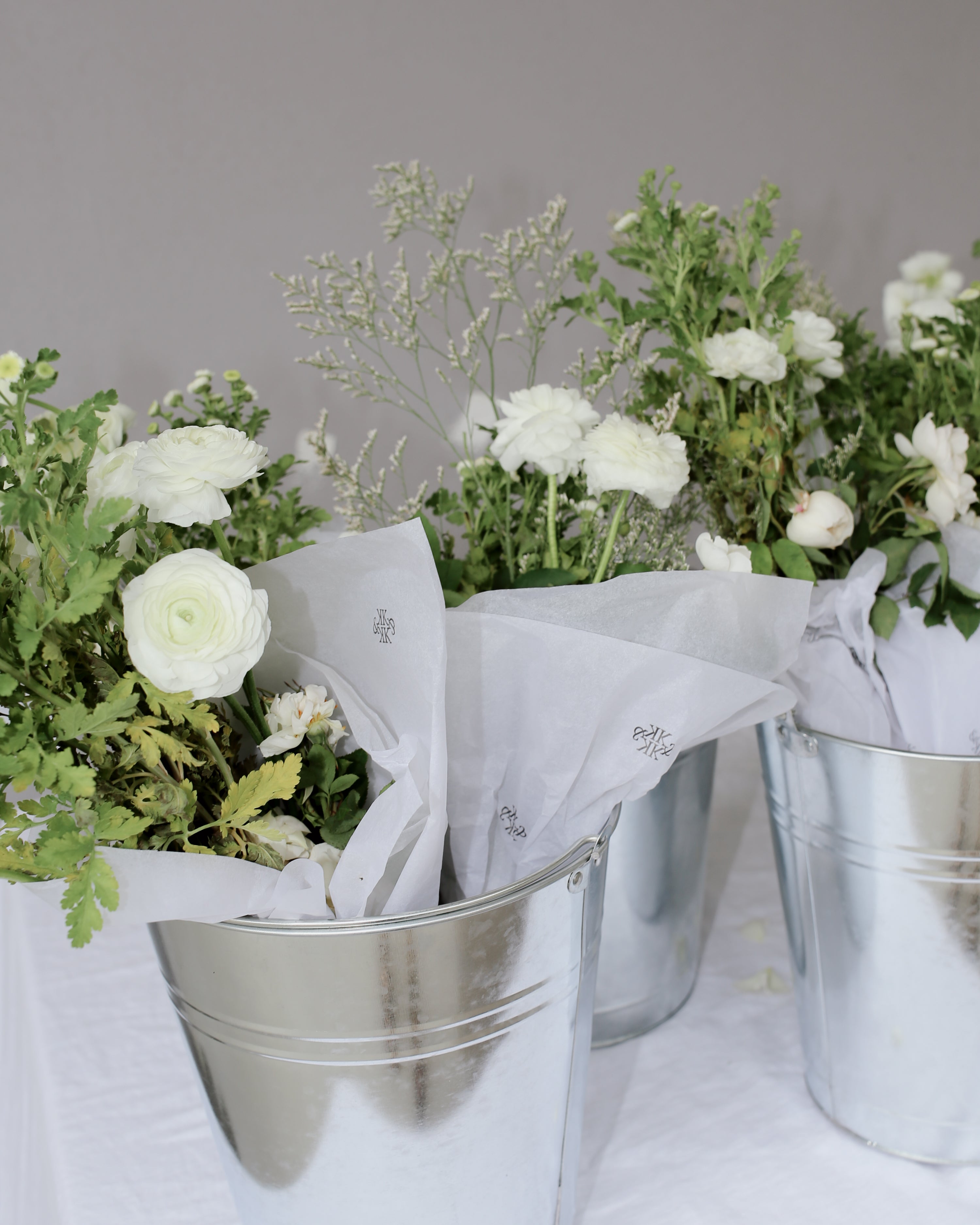 Silver buckets with white roses and greenery wrapped in tissue paper, set on a white tablecloth, exuding high-end floral elegance.