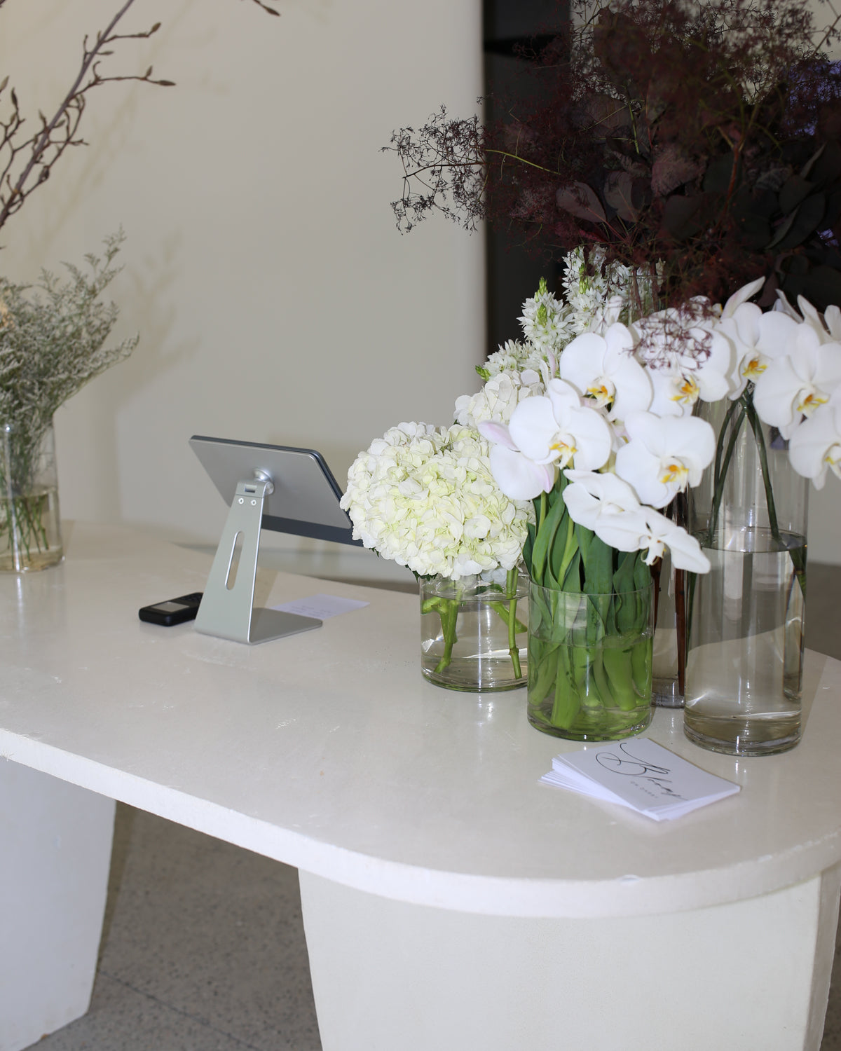 Luxurious jewellery store counter adorned with elegant white orchids, hydrangeas, and earthy foliage in glass vases, alongside digital tablet display.