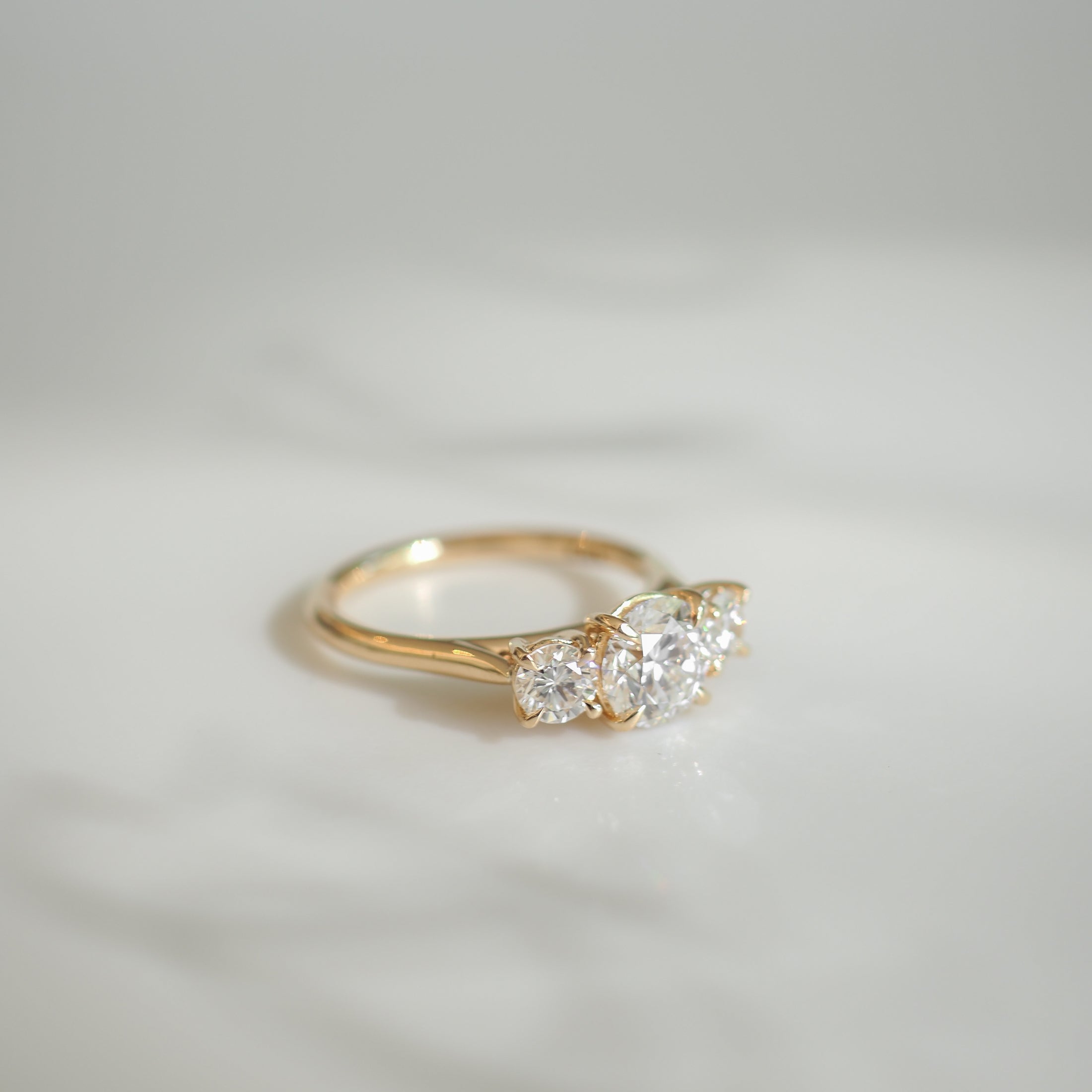 Elegant gold ring featuring three sparkling diamonds, set against a soft, blurred white background, exuding luxury and sophistication.