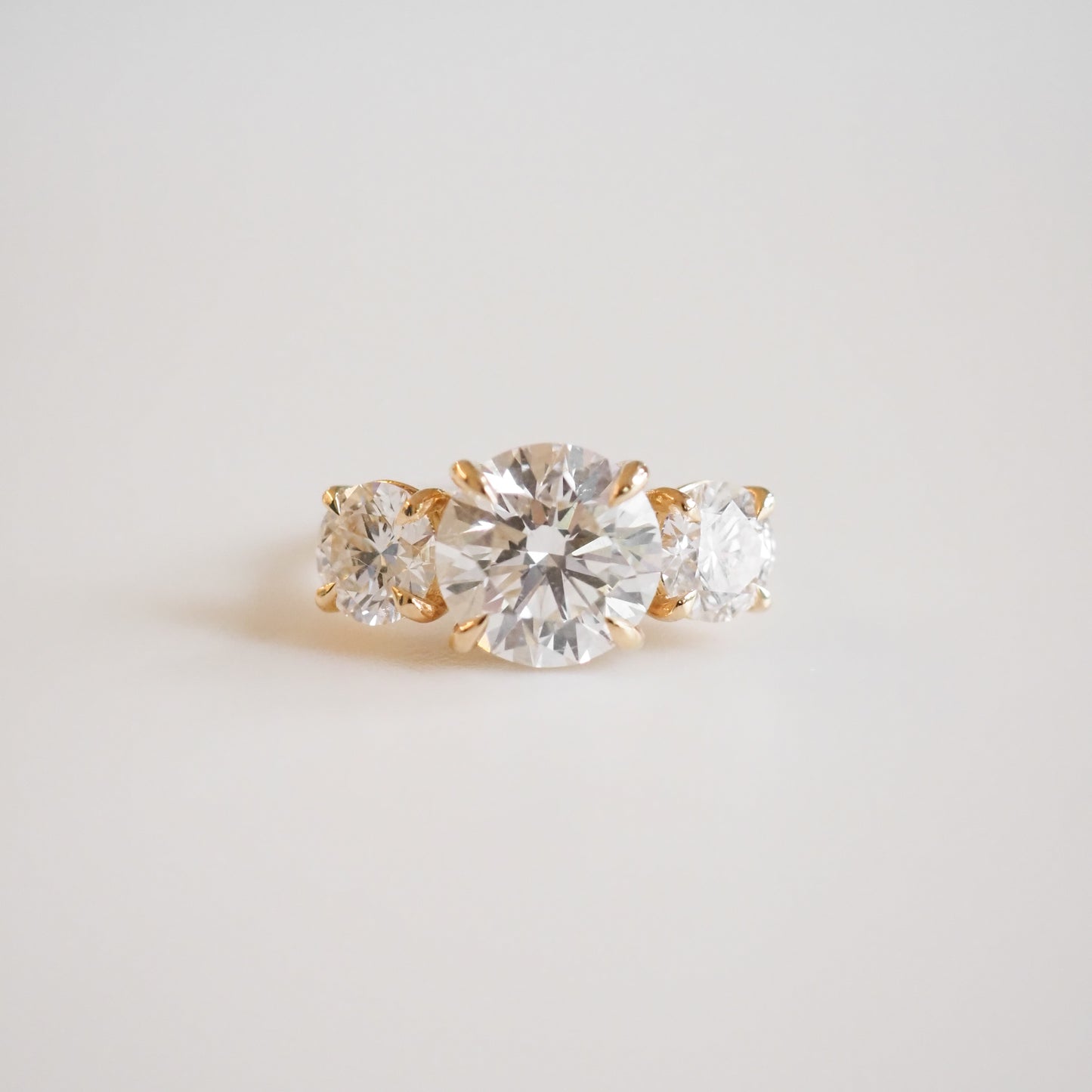 Elegant gold engagement ring with three brilliant round-cut diamonds, centred by a larger diamond, on a soft white background. Luxurious jewellery.