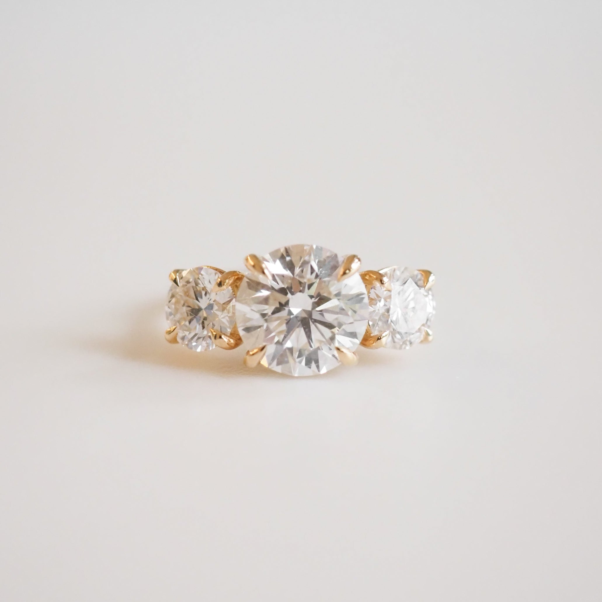 Elegant gold engagement ring with three brilliant round-cut diamonds, centred by a larger diamond, on a soft white background. Luxurious jewellery.