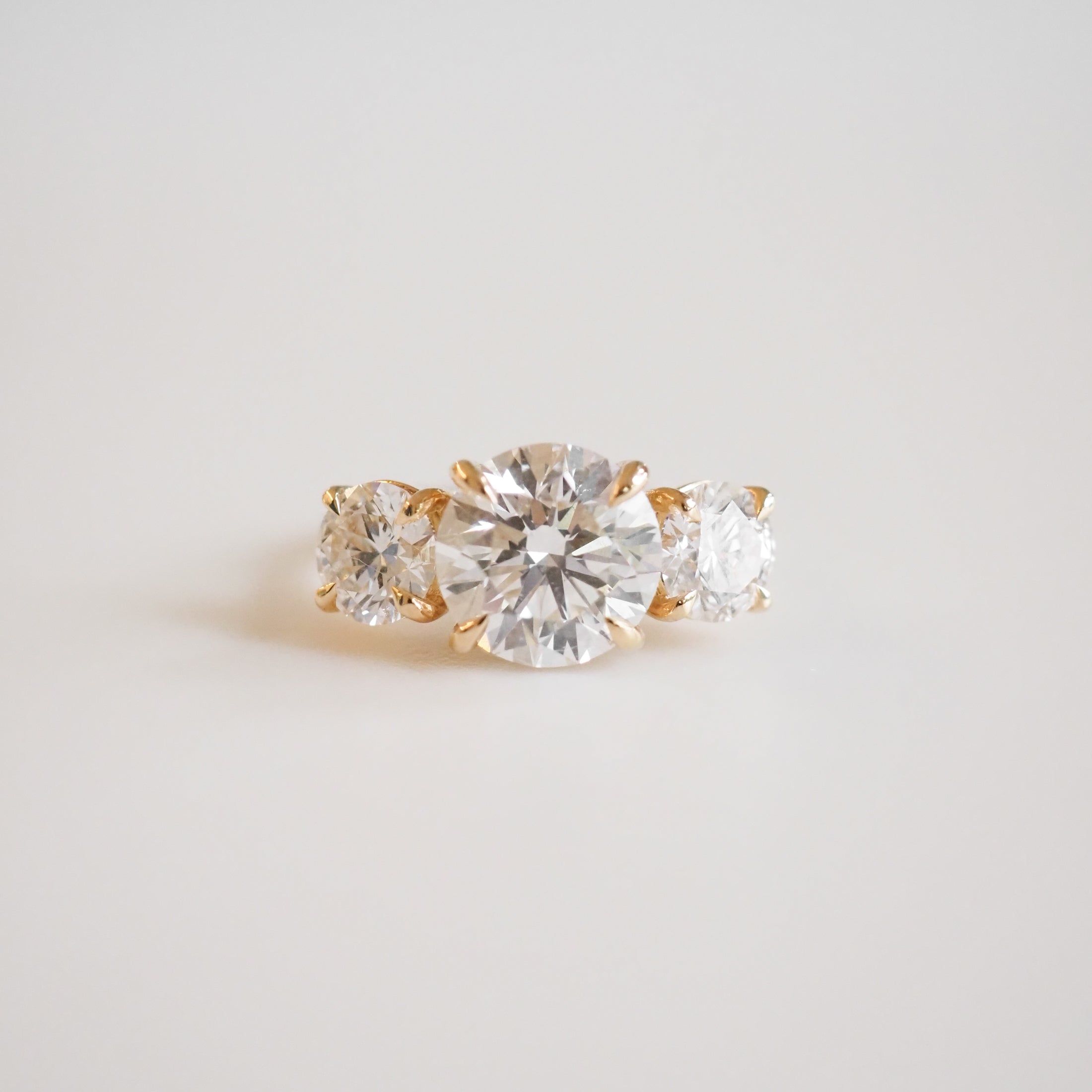 Elegant gold engagement ring with three brilliant round-cut diamonds, centred by a larger diamond, on a soft white background. Luxurious jewellery.
