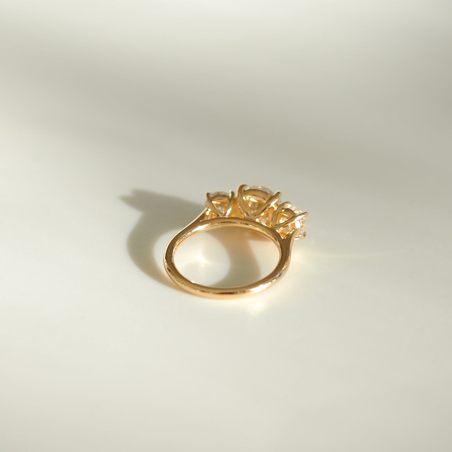 Gold ring with intricate triple stone setting on a light background, casting a soft shadow. Luxurious fine jewellery piece.