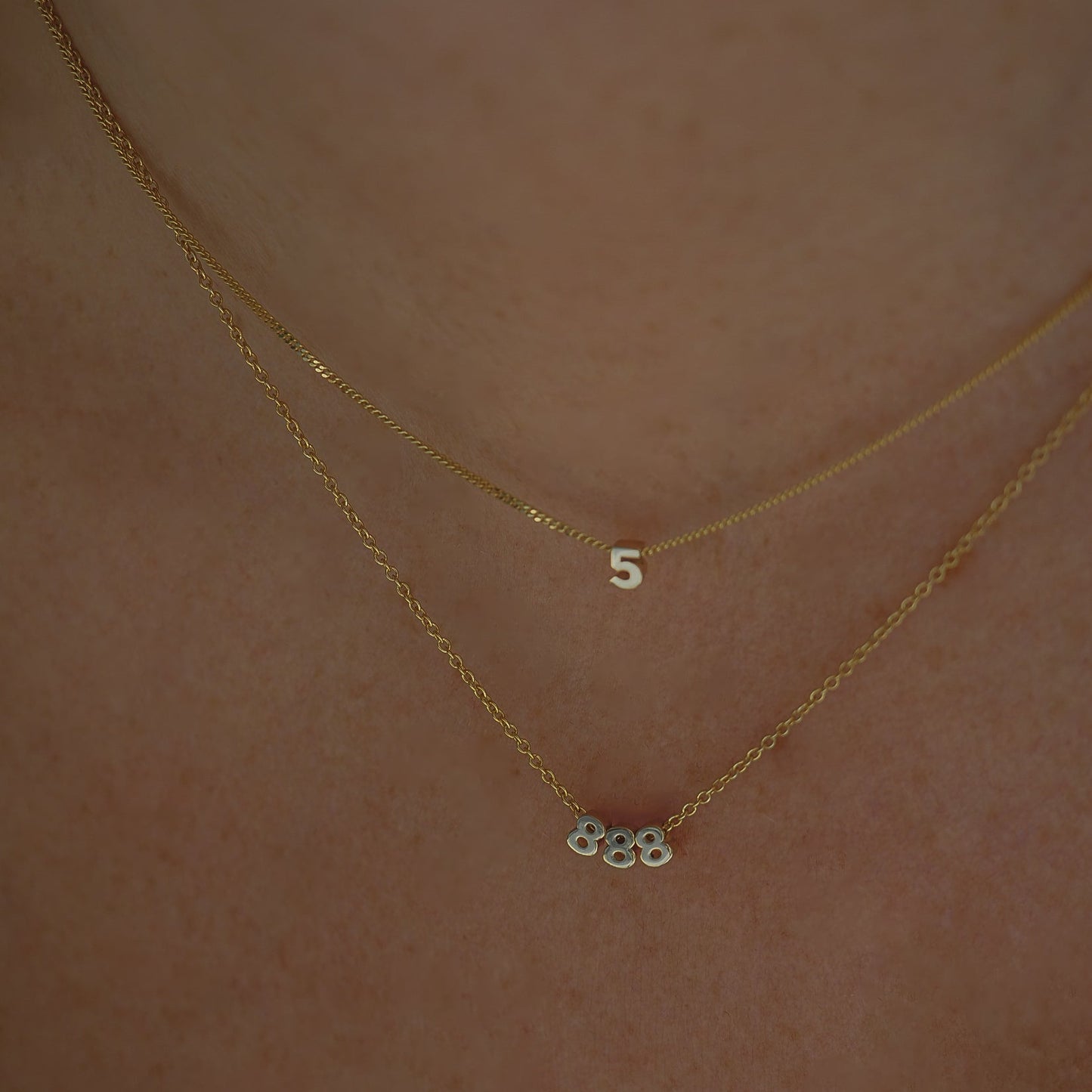 Two gold necklaces, one with a numeral "5" pendant and the other with "888," on a person's neck. Elegant, luxury jewellery.