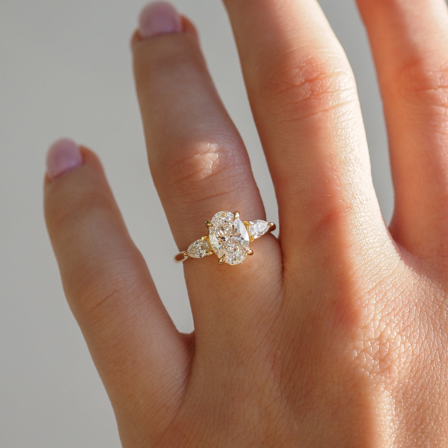 Close-up of a gold ring with an oval diamond centre and pear-shaped side stones, worn on a hand with light pink nails.