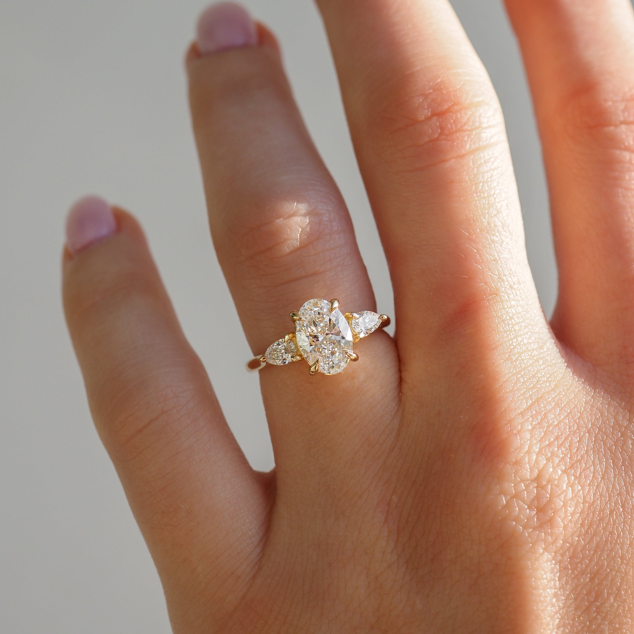 Close-up of a gold ring with an oval diamond centre and pear-shaped side stones, worn on a hand with light pink nails.