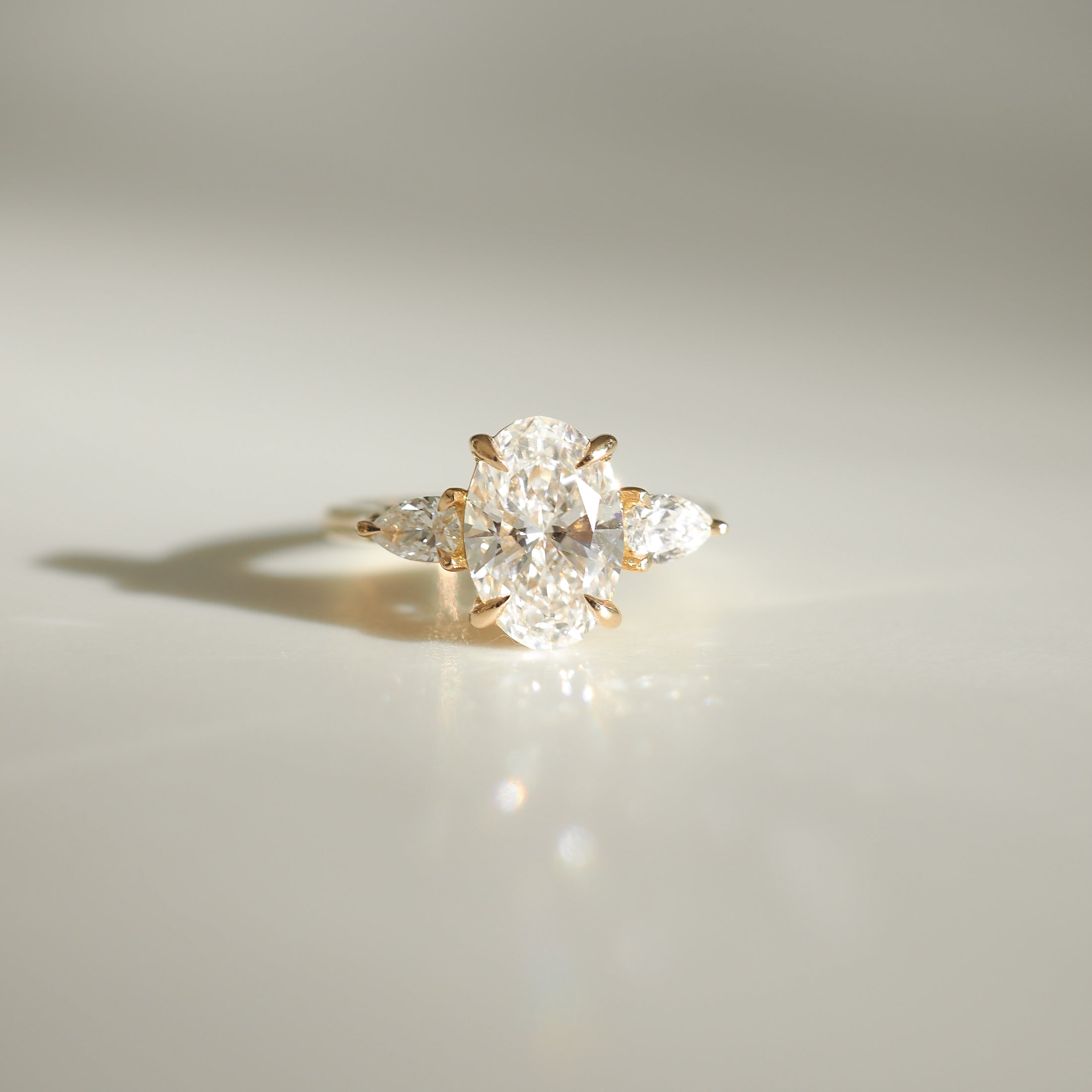 Elegant gold ring with a central oval diamond flanked by two pear-shaped diamonds on a smooth reflective surface. Luxurious jewellery.