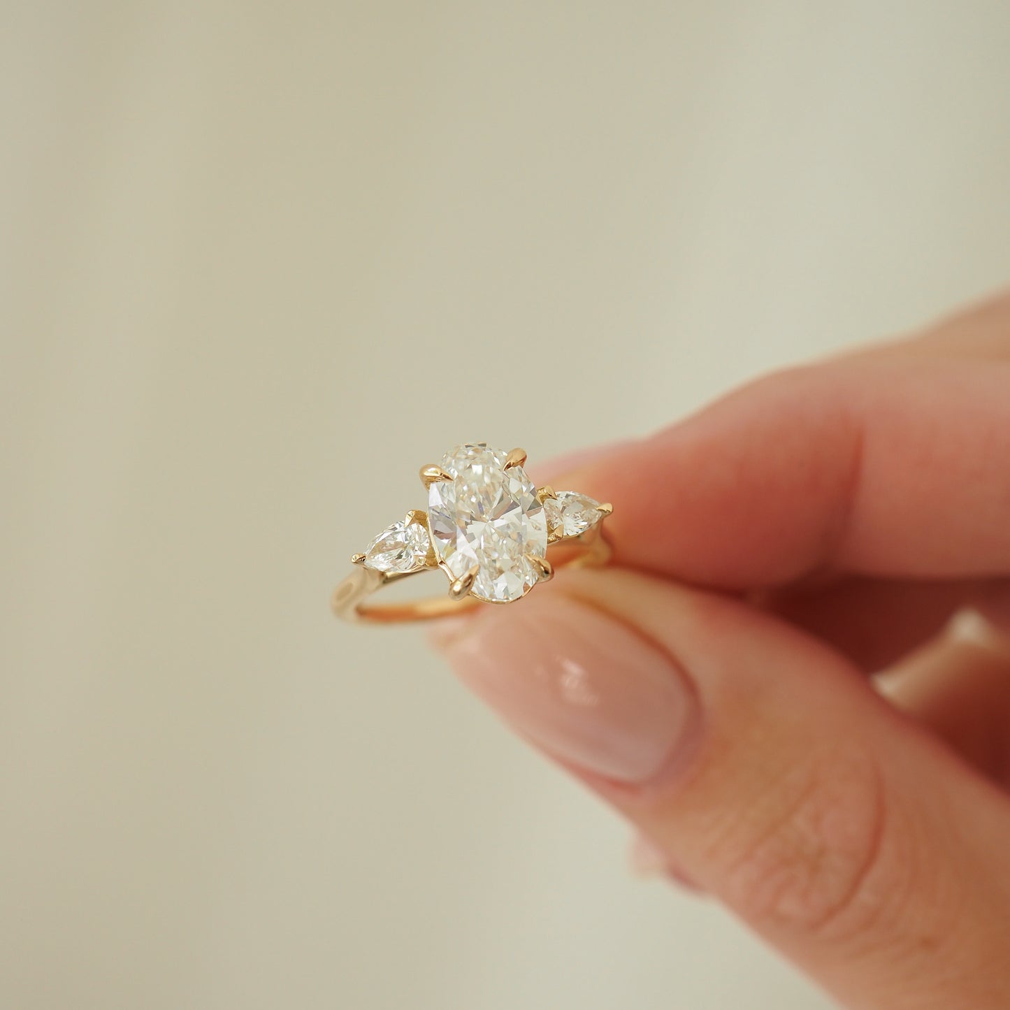 Hand holding an elegant gold ring with a large oval diamond flanked by two smaller pear-shaped diamonds, set against a soft background.
