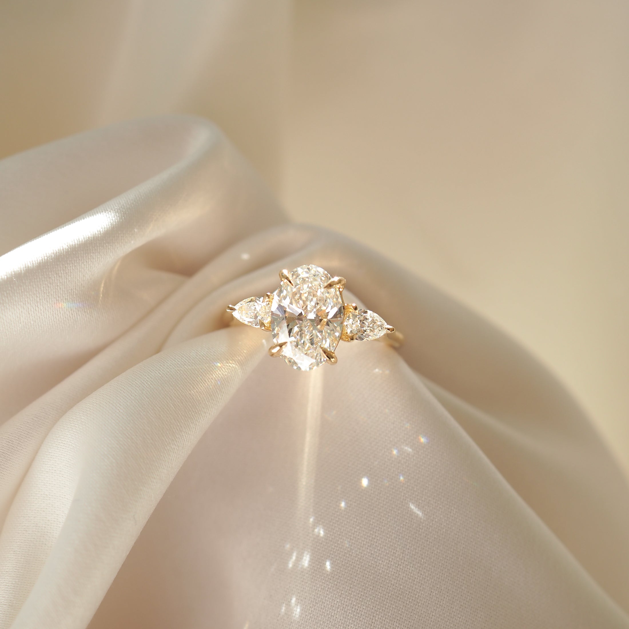 Elegant gold ring with a large oval diamond and two pear-shaped diamonds, resting on a silky fabric. Luxurious jewellery display.