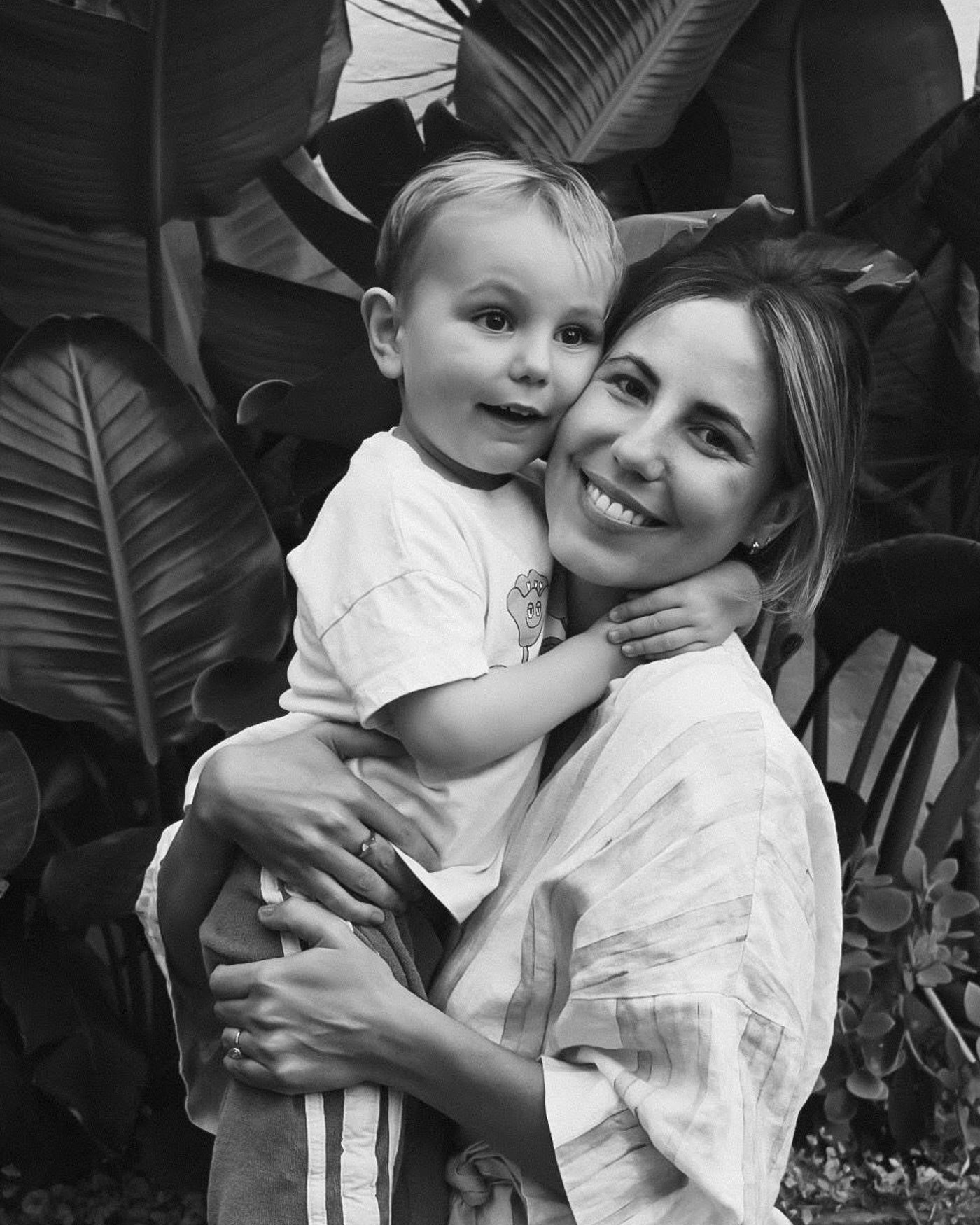 A woman wearing a ring holds a smiling child in front of lush tropical leaves, showcasing family joy and luxury jewellery. Monochrome photo.