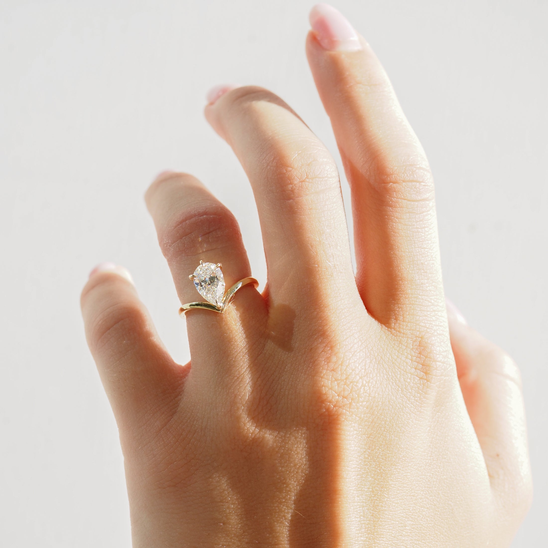 A hand wearing an elegant gold ring with a large, pear-shaped diamond against a neutral background, showcasing luxury jewellery design.