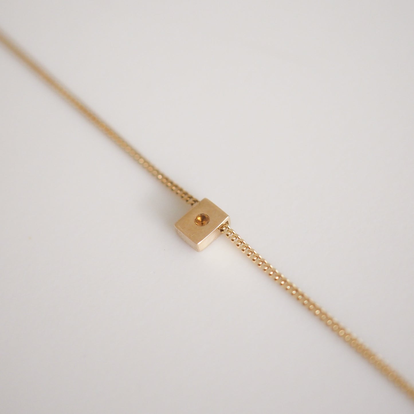 Gold chain necklace with a minimalist square pendant featuring a central bead on a white background. Luxurious fine jewellery design.