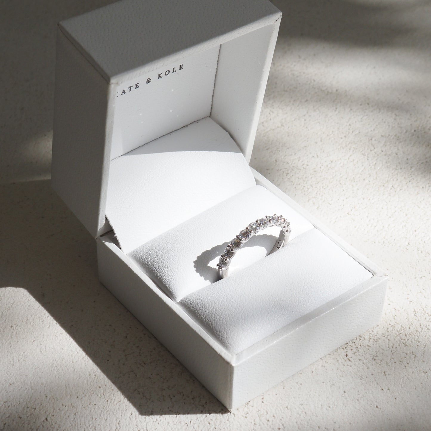 A sparkling diamond ring in a white luxury gift box from Kate & Kole, set against a textured, softly lit background.