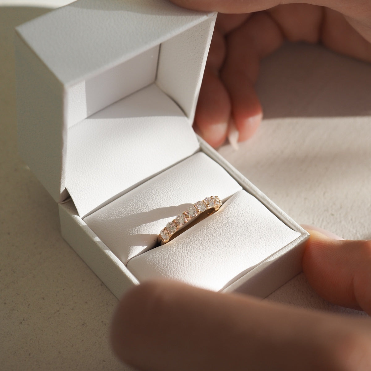 Elegant gold ring with sparkling gemstones in an open white jewellery box, hands holding the box under soft lighting.