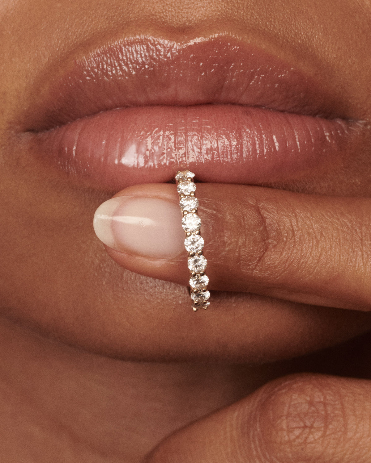A close-up of a person's lips and fingers wearing a thin gold band ring set with clear round-cut diamond.