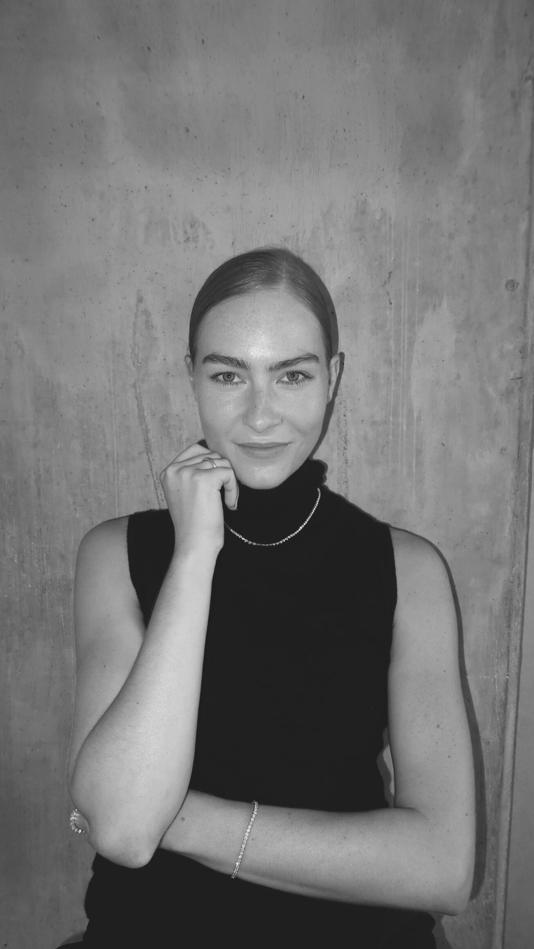 A person in a sleeveless black top, wearing a delicate pearl necklace and bracelet, poses confidently against a textured concrete wall.