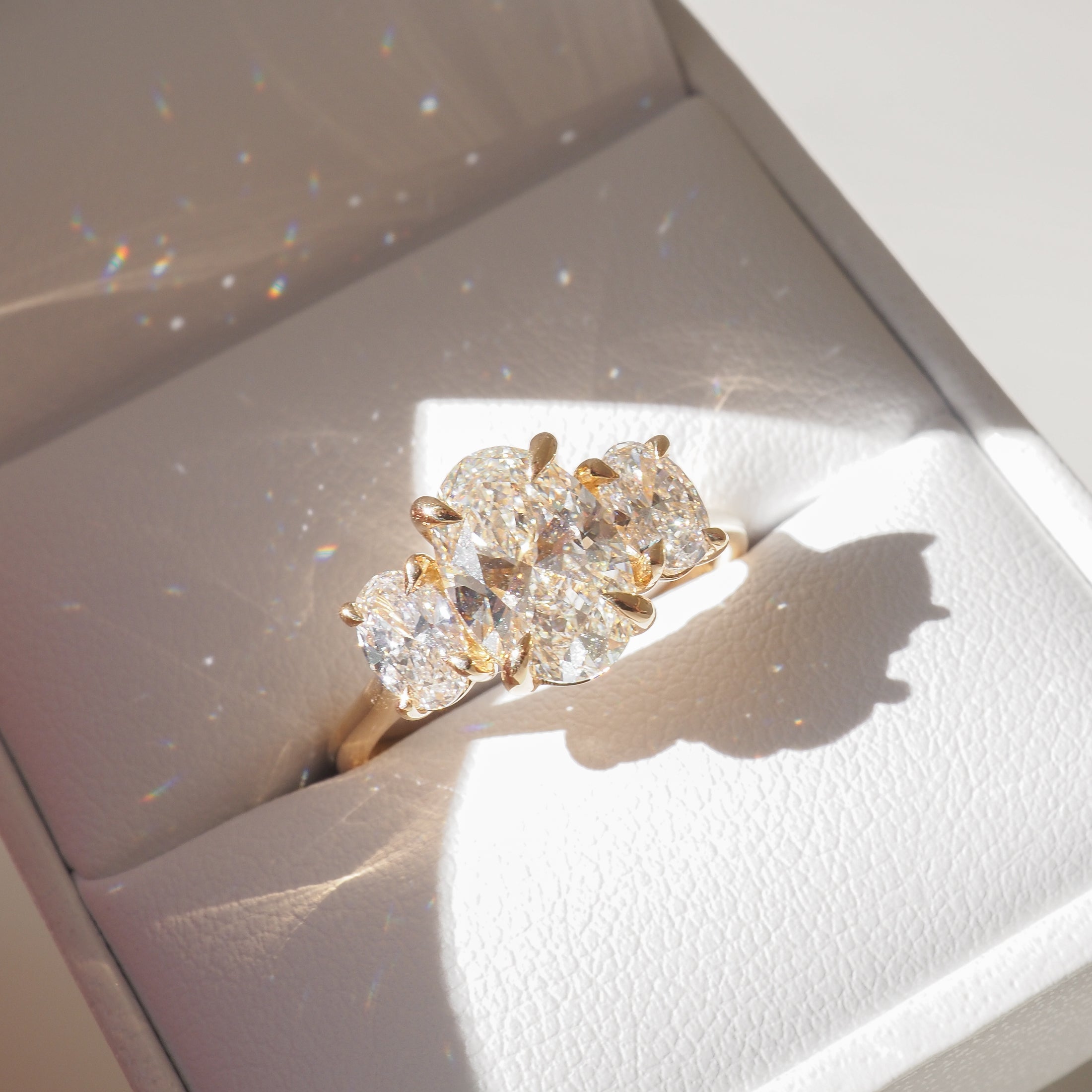 Gold ring with three large oval-cut diamonds in a white jewellery box, sparkling under soft light. Perfect for luxury engagement or bridal jewellery.