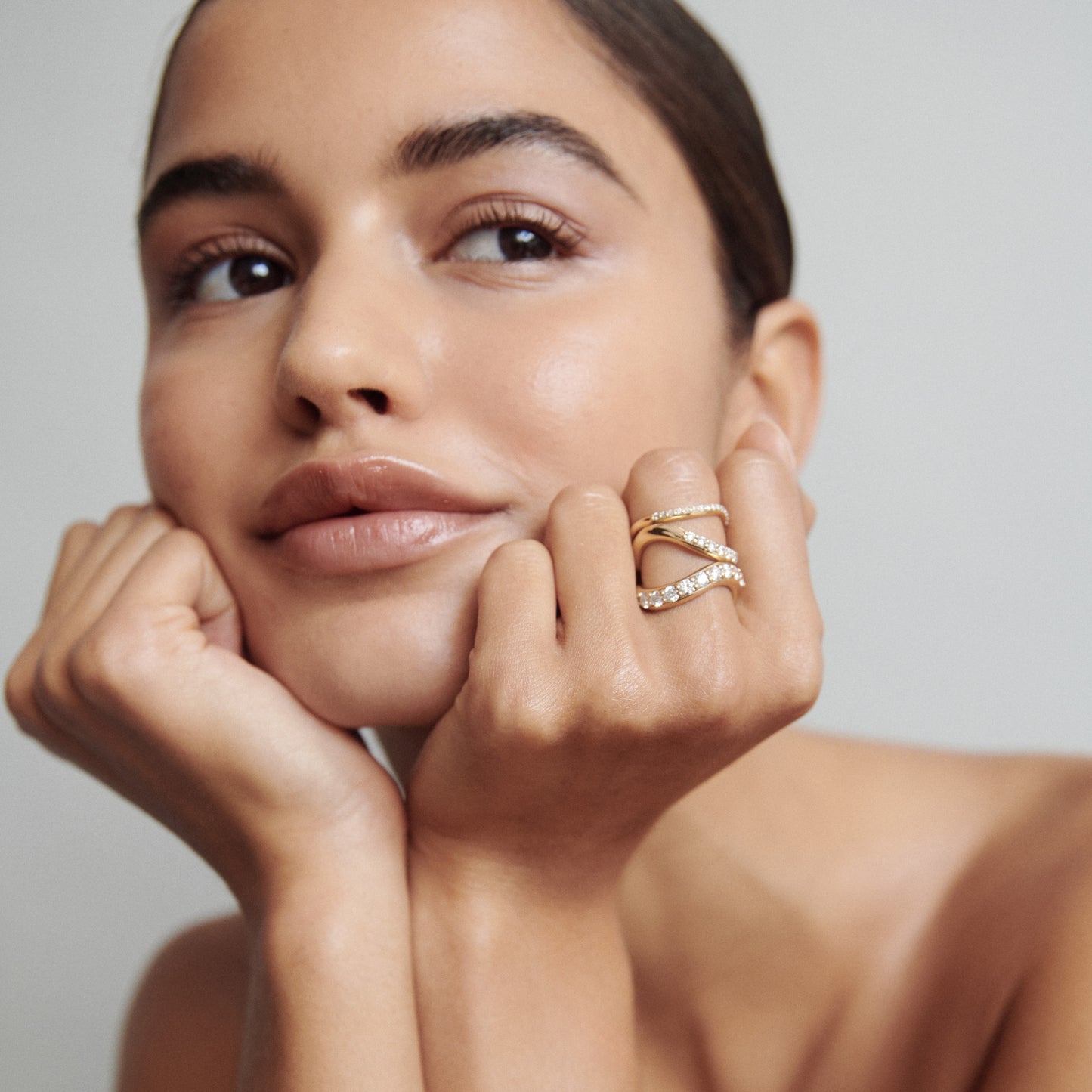 Woman with a serene expression wearing a gold and diamond criss-cross ring, resting her chin on her hands against a light grey background.