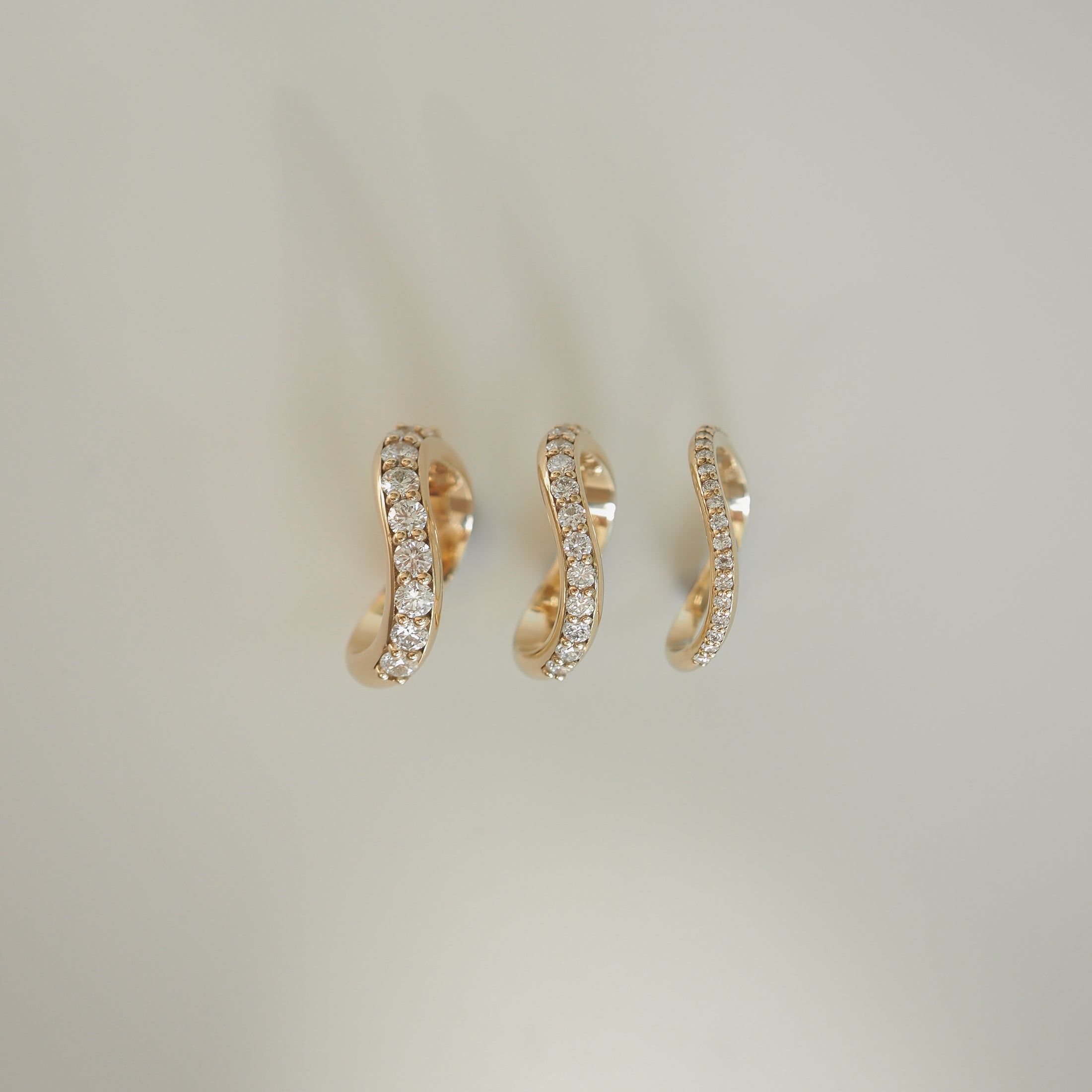Three elegant, gold wavy hoop earrings adorned with diamonds, displayed against a soft, neutral background. Luxurious fine jewellery.