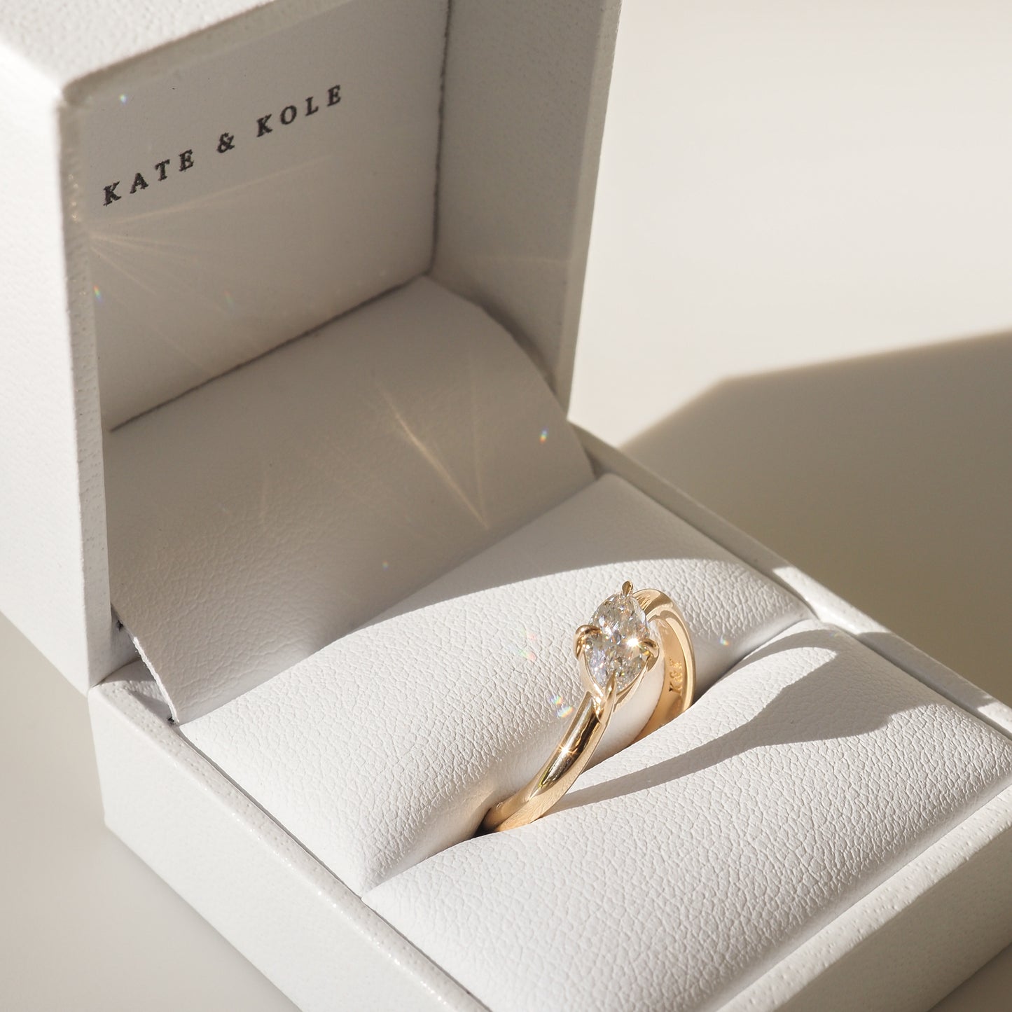 Gold engagement ring with a diamond, elegantly displayed in a white Kate & Kole jewellery box, casting a soft shadow.