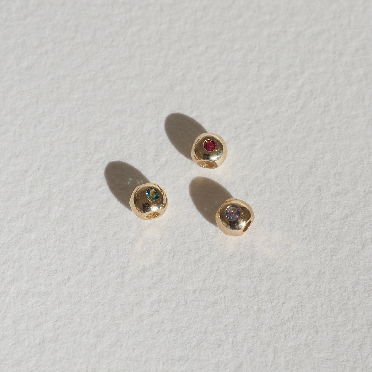 Three small gold stud earrings with red, green, and grey gemstones on a textured white surface, casting soft shadows.