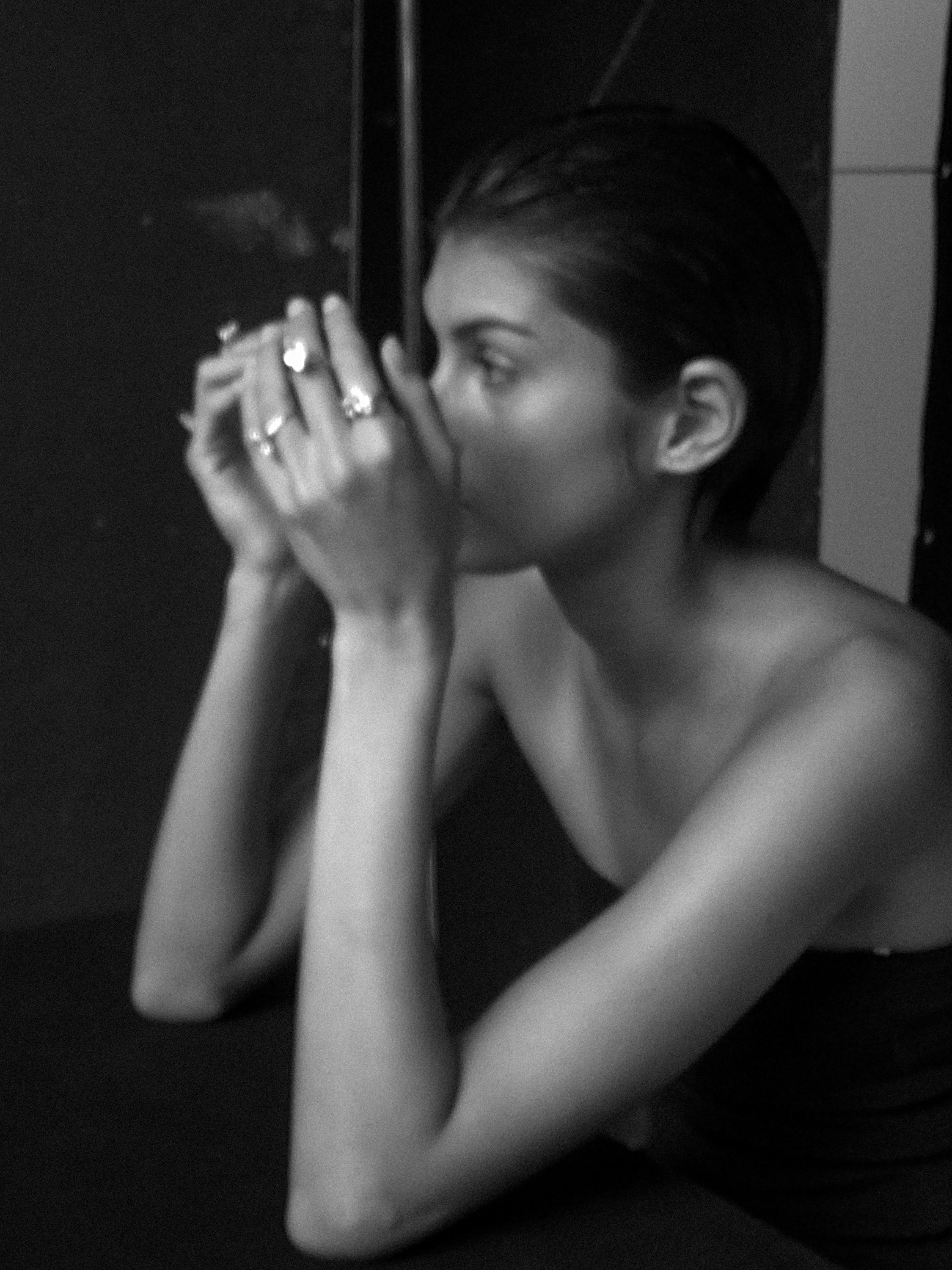 Black and white image of a woman in a strapless top, showcasing luxury rings on both hands, holding an object close, with a pensive expression.
